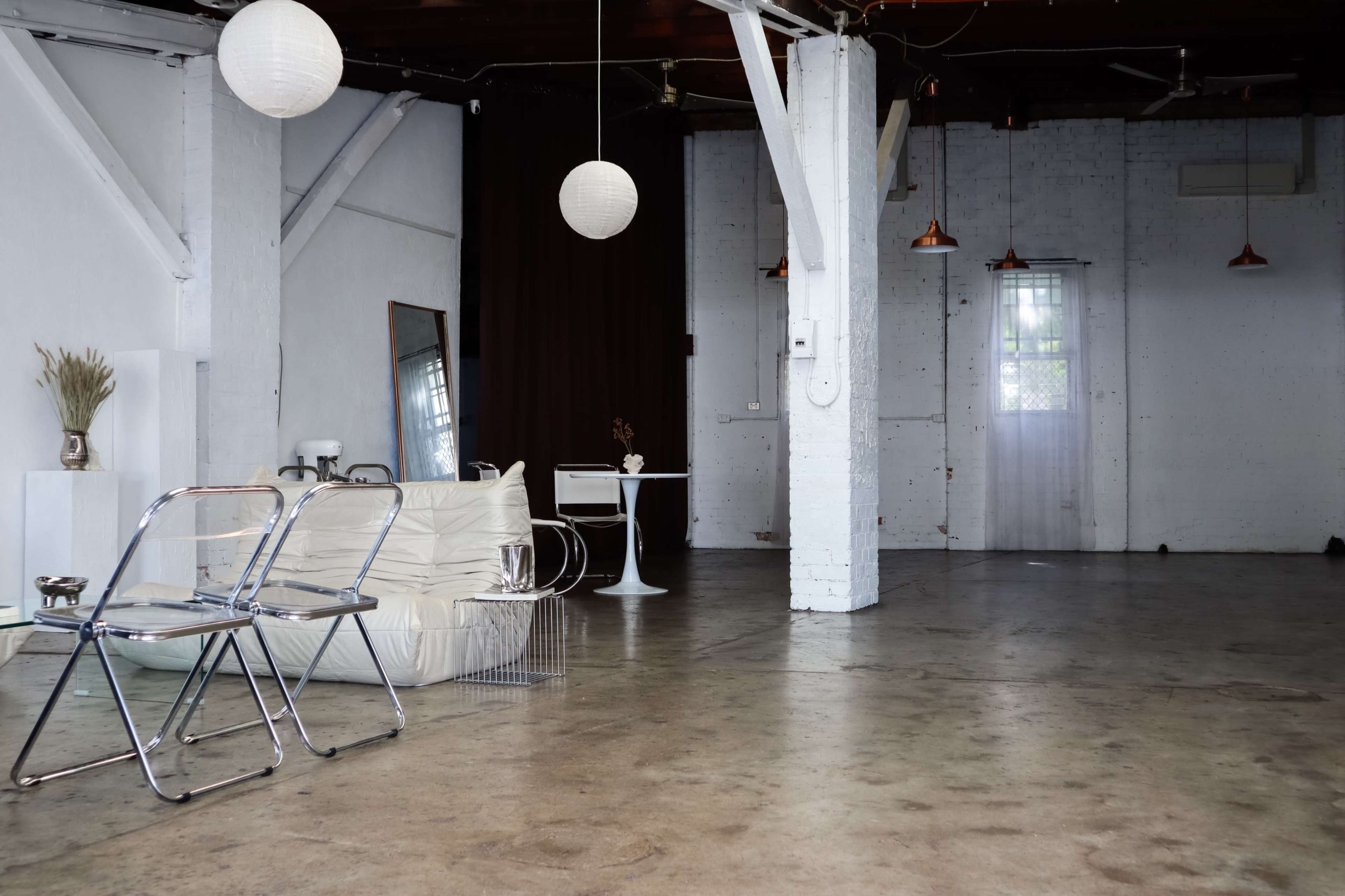 The image shows a spacious, minimalist interior with white walls, concrete flooring, and various furniture pieces including a table and chairs.