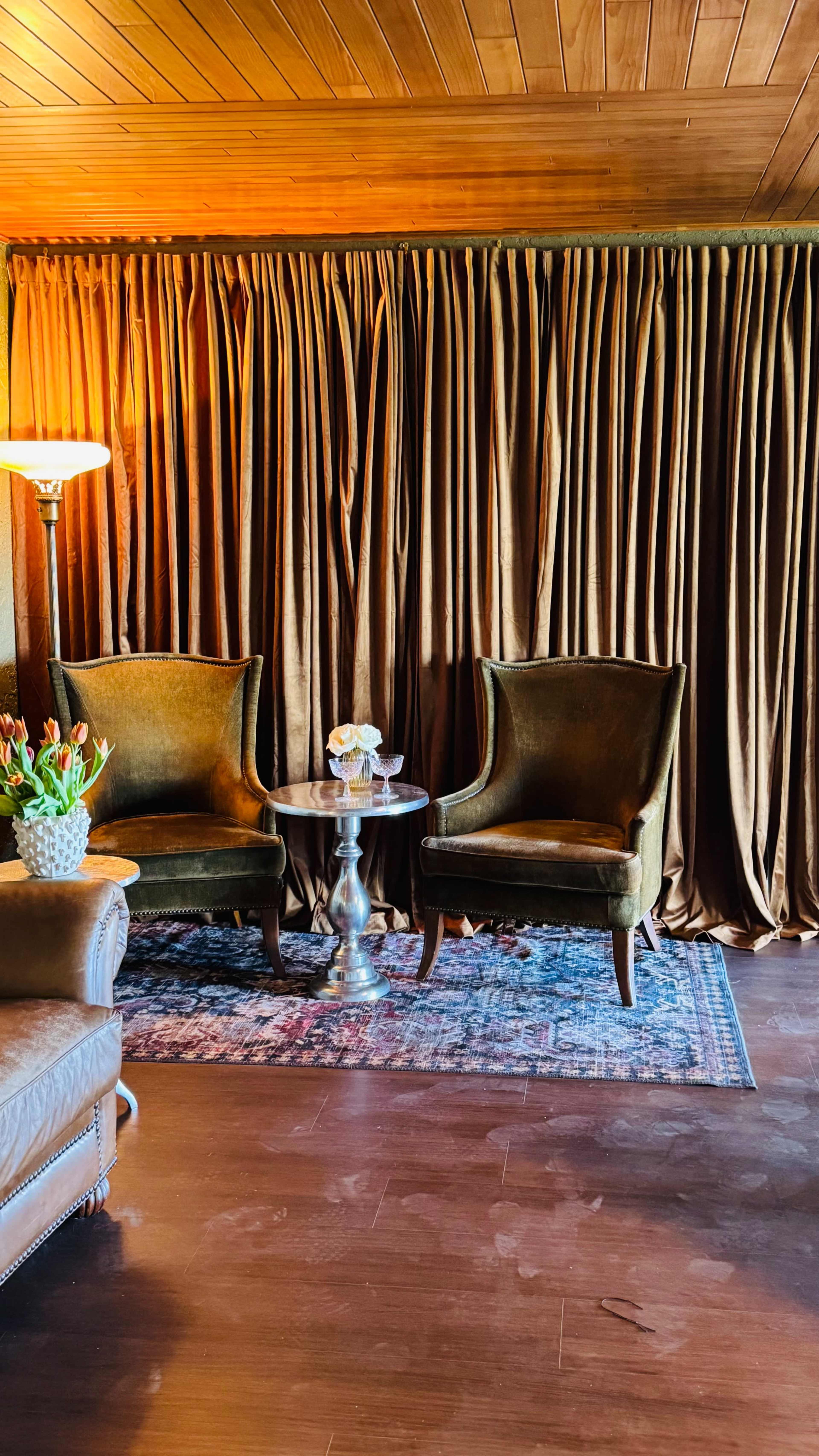 The image shows a cozy seating area with two green armchairs flanking a small table, set against a backdrop of floor-length brown curtains.