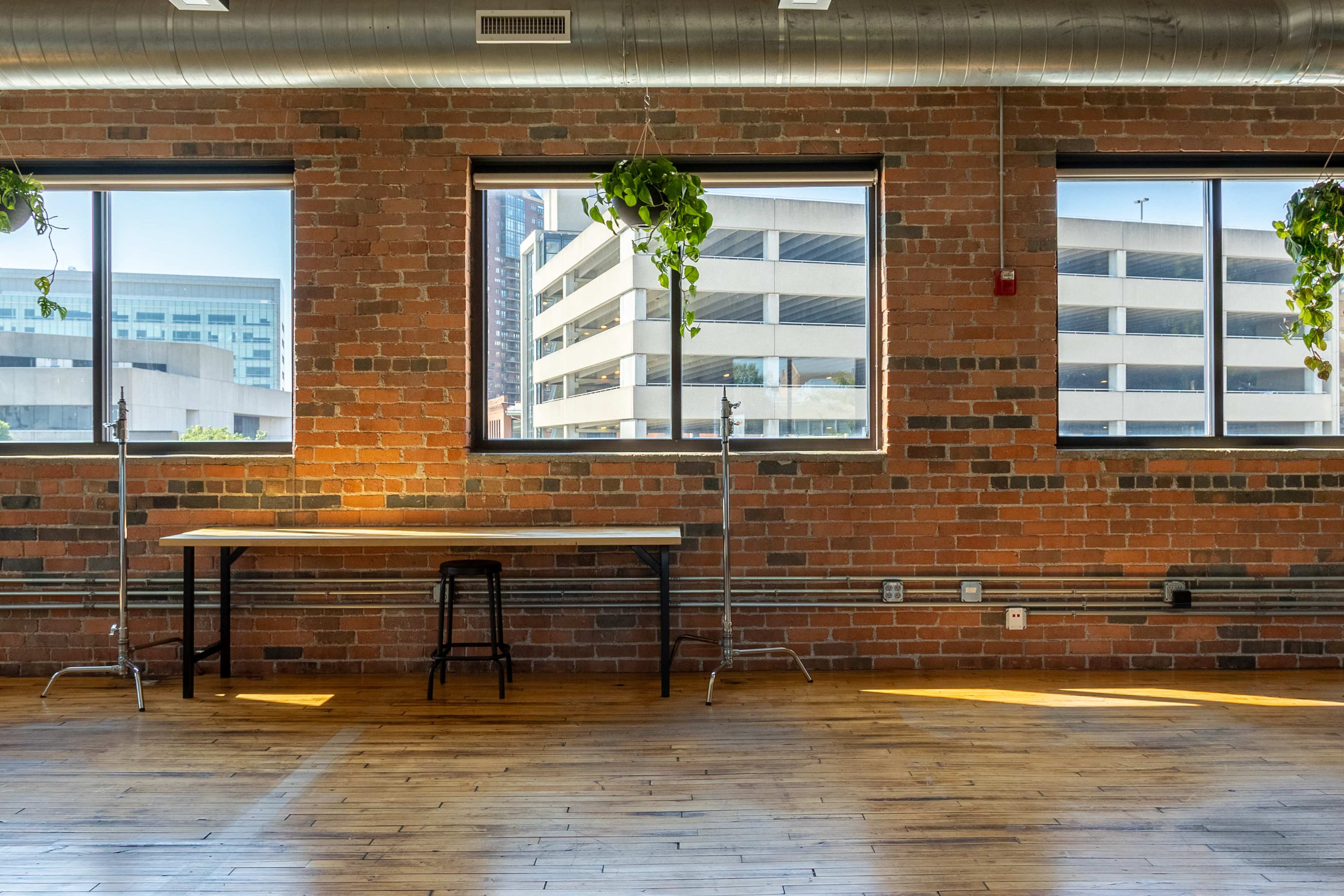 A large, open room with wooden flooring, exposed brick walls, and large windows overlooking a parking structure, featuring a table and a stool positioned near the windows.