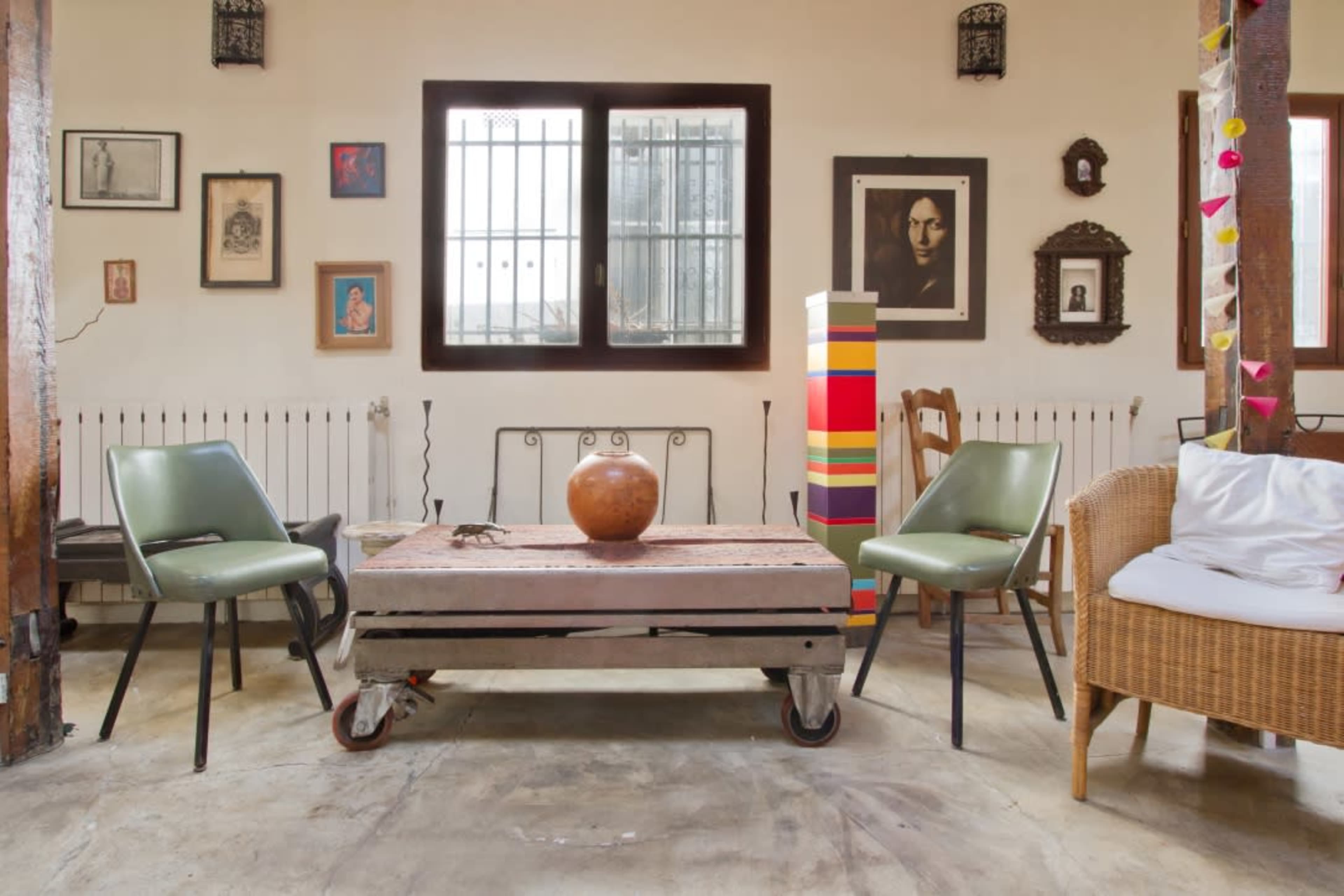 A cozy interior with a wooden table on wheels, two green chairs, a decorative bowl, and various framed artworks on the walls.