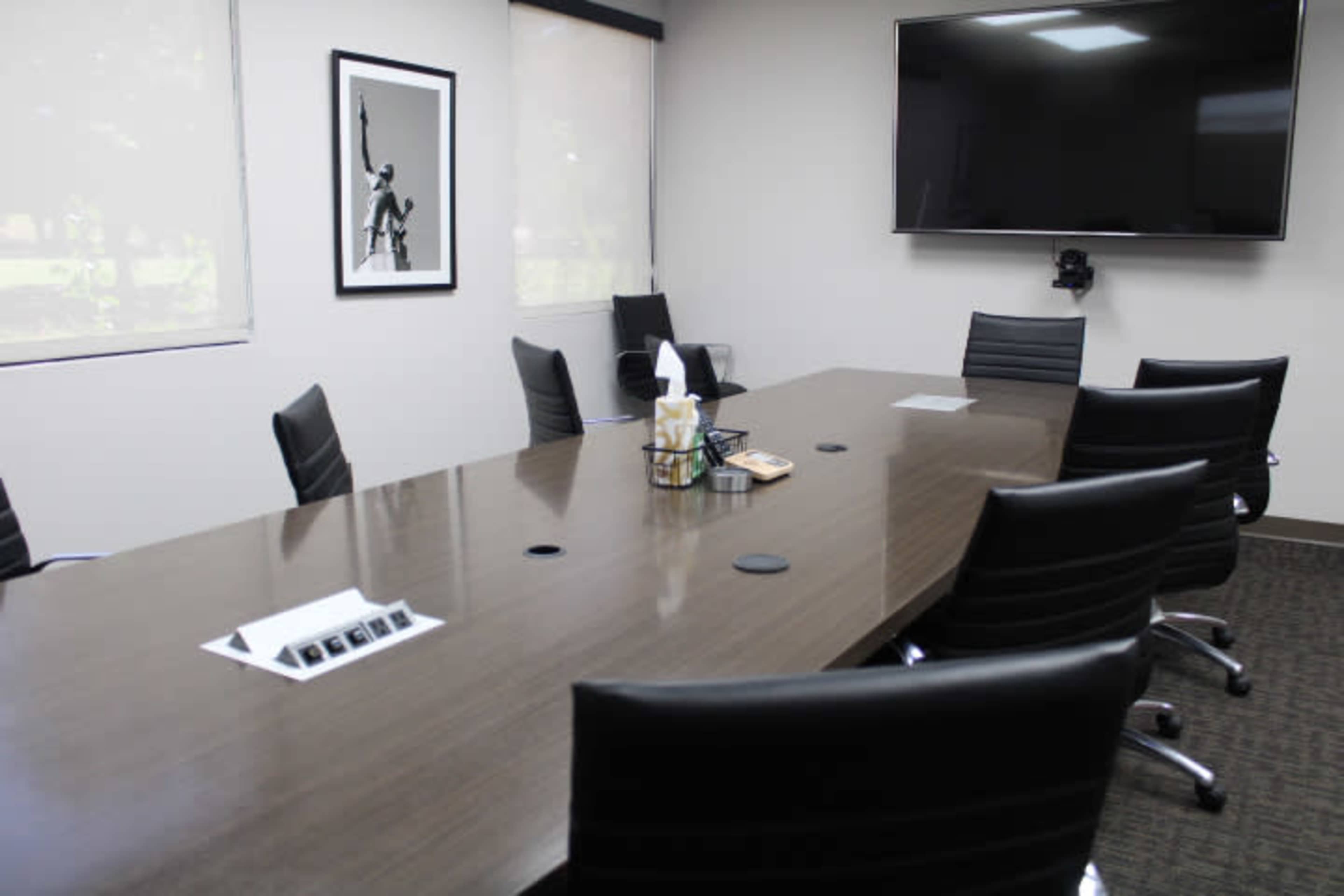 The image shows a modern conference room with a long wooden table, several black chairs, and a large wall-mounted screen.
