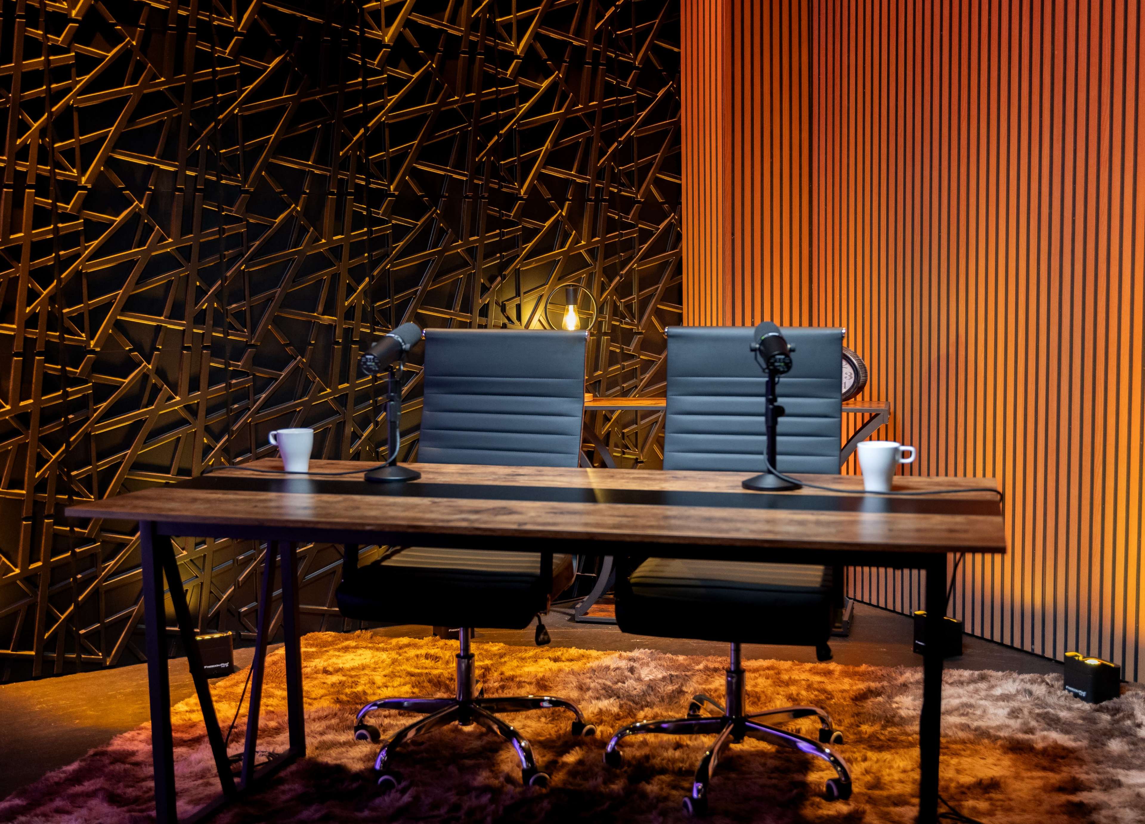 The image shows a modern studio setup featuring two black leather chairs with microphones on a wooden table, set against a backdrop of textured wall panels illuminated with warm lighting.