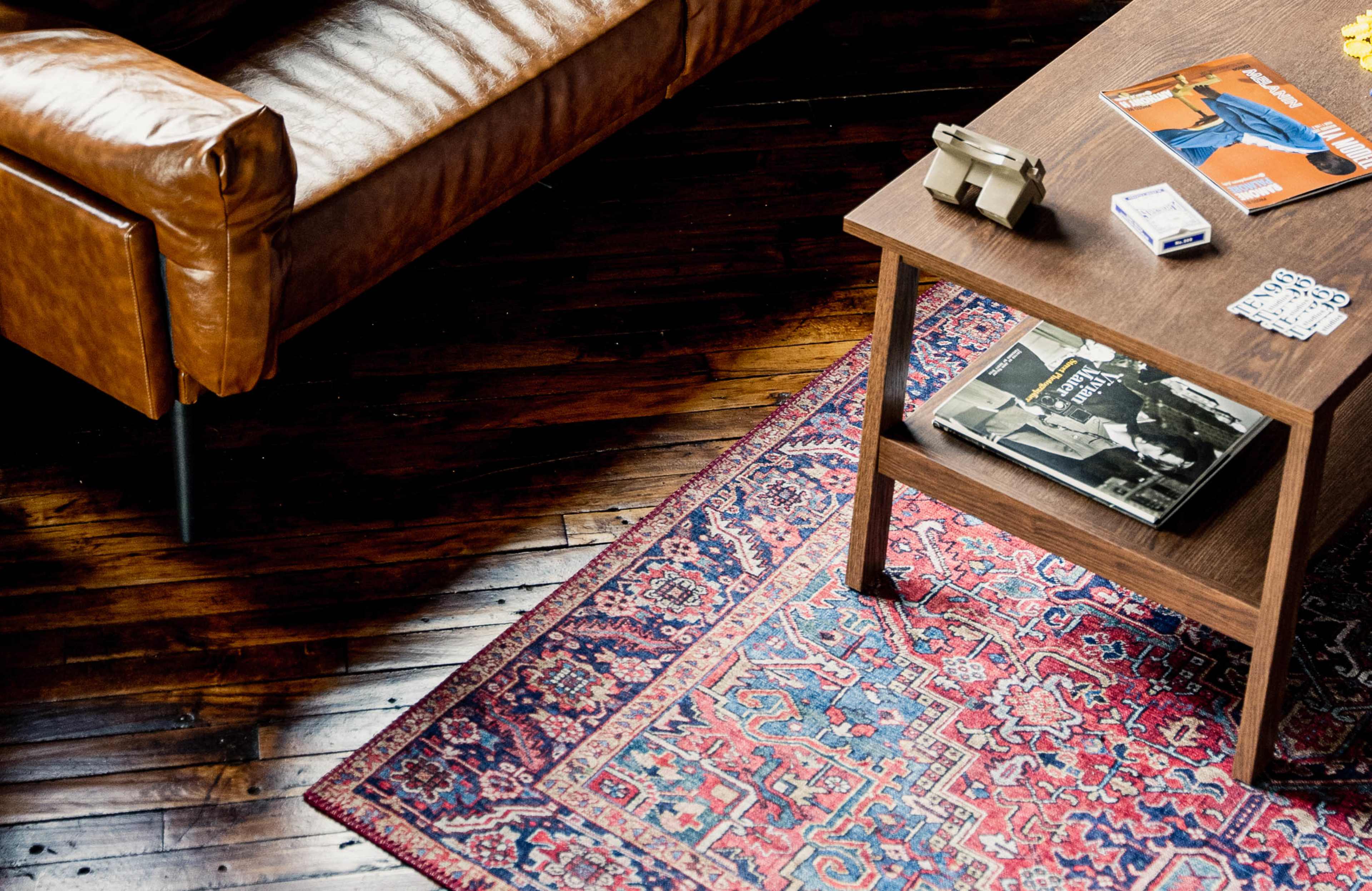 A brown leather sofa sits beside a wooden coffee table adorned with magazines and a vintage camera, placed on a patterned rug over a wooden floor.