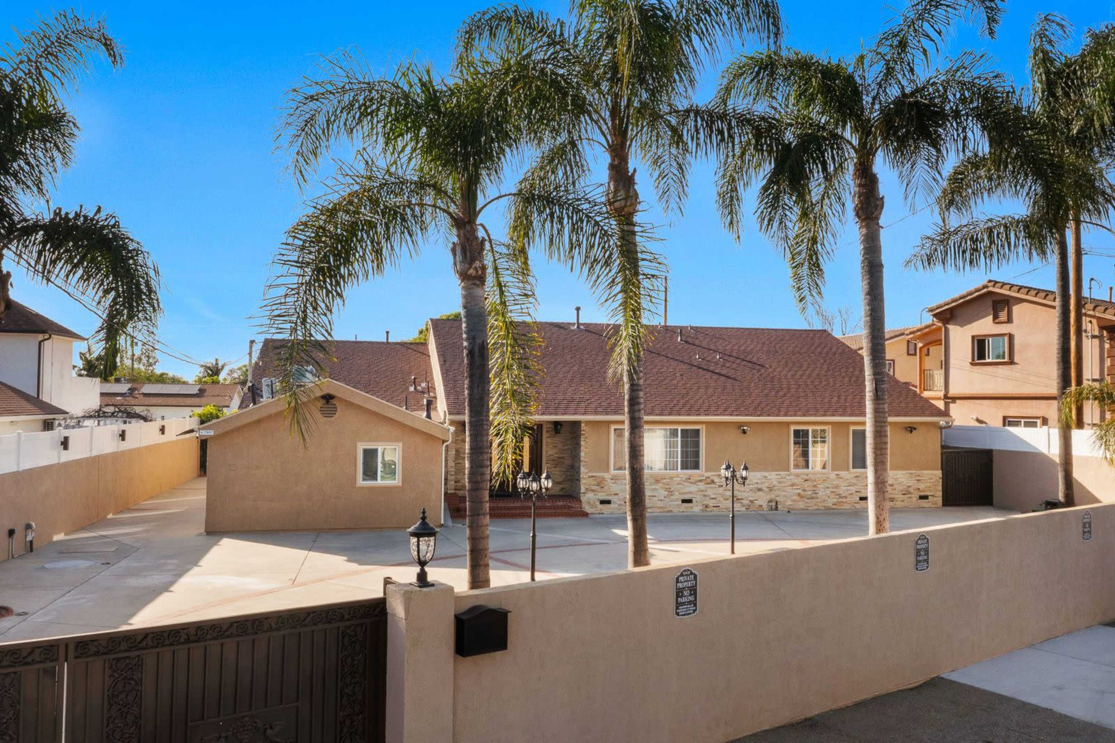 The image shows a single-story house surrounded by palm trees and a large, enclosed driveway.