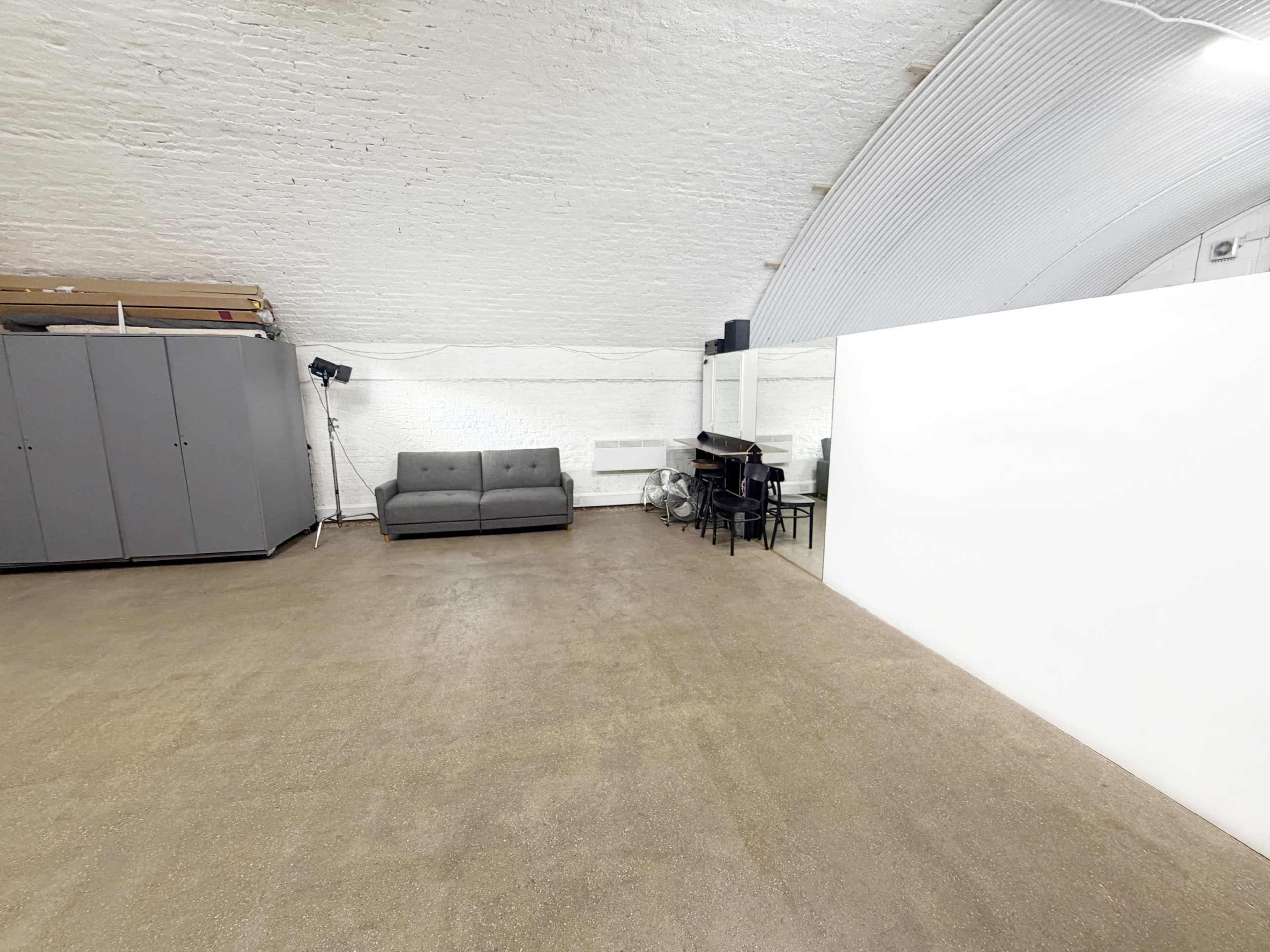 The image shows a spacious, empty interior with a gray couch on one side and minimalist furniture, including a table and chairs, on the opposite side against a white wall.