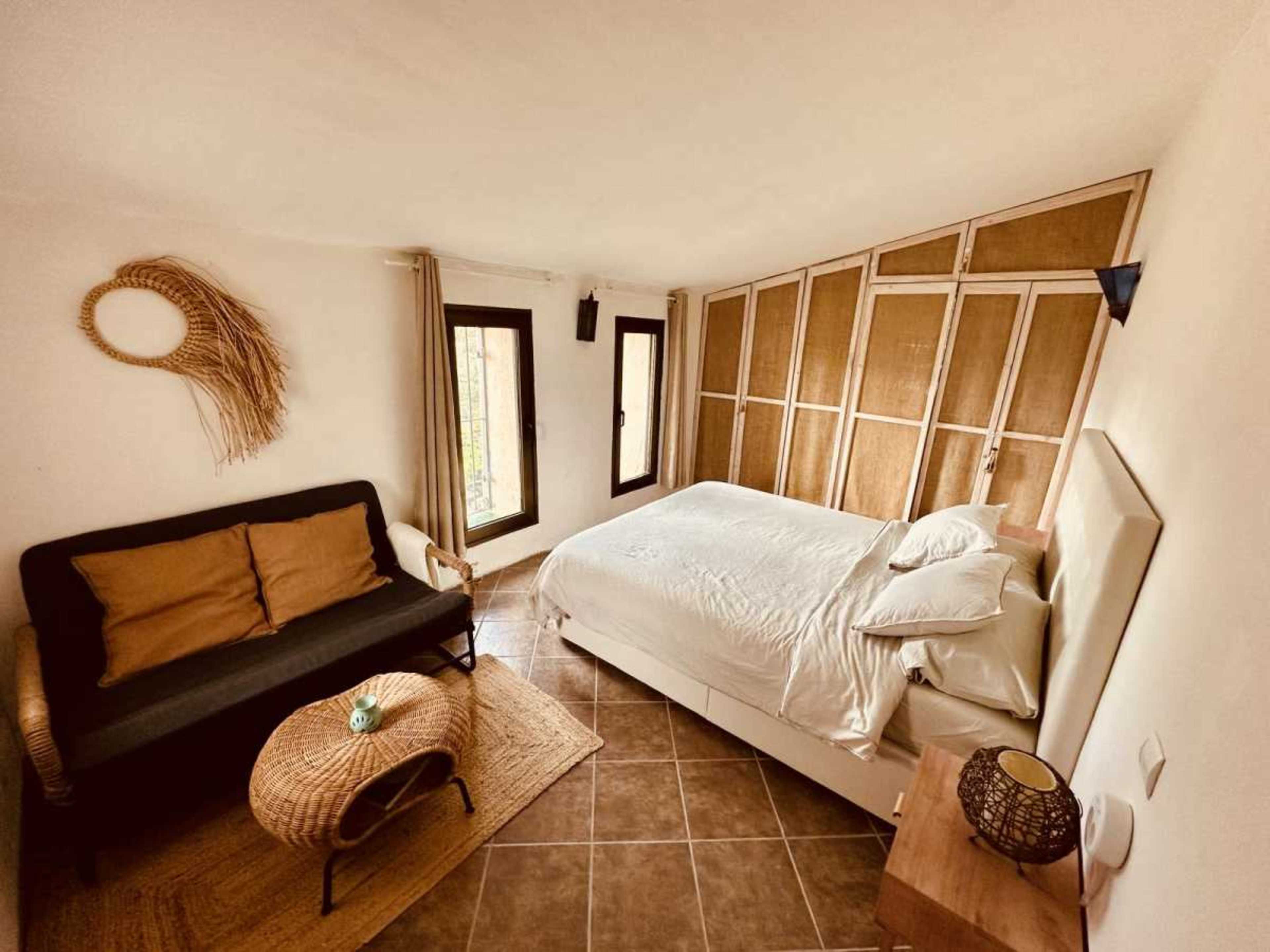 The image shows a minimalist bedroom featuring a double bed, a black sofa, and a round coffee table, with natural light streaming through large windows.
