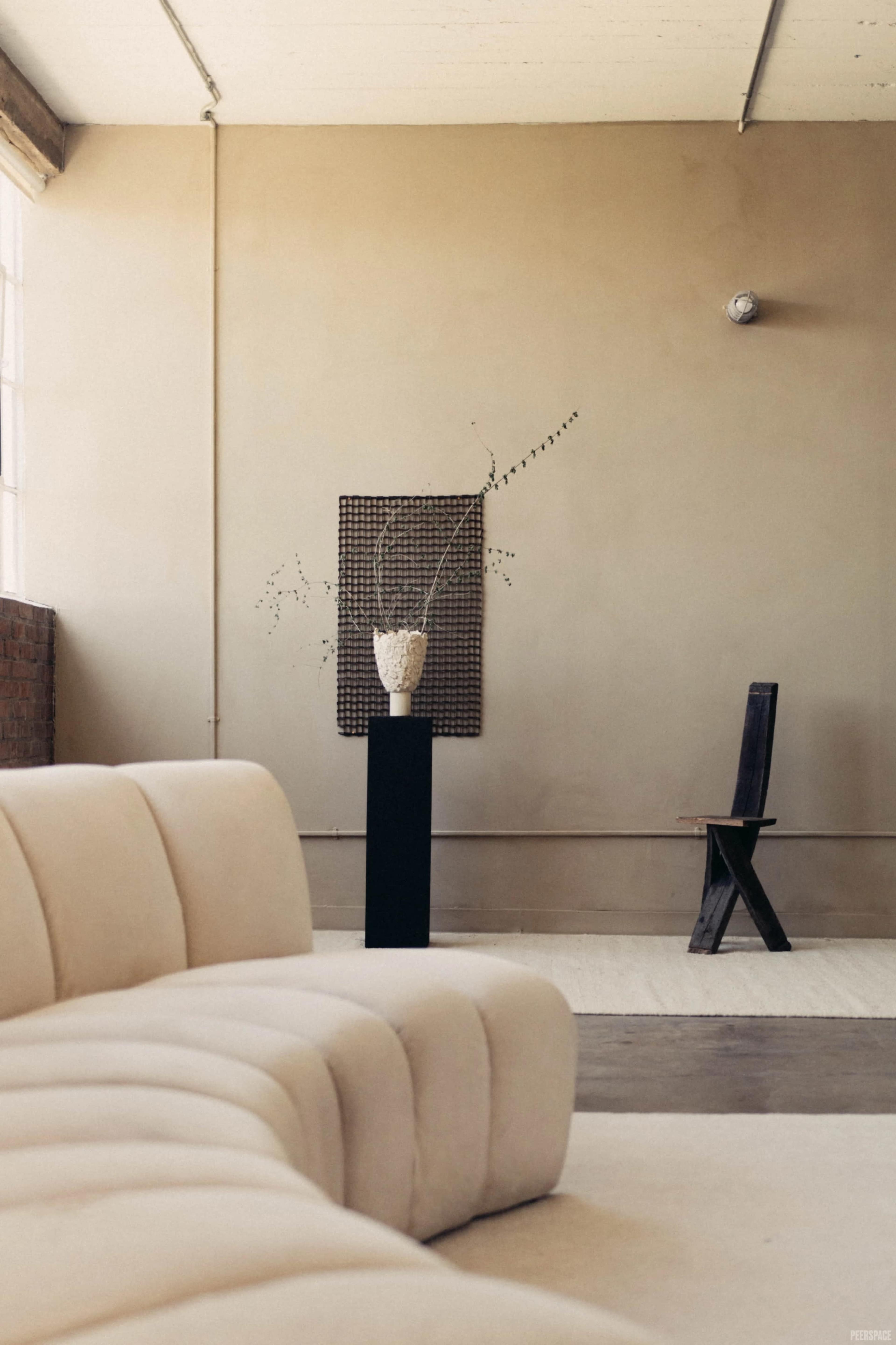 A beige, curved sofa sits in the foreground, with a textured vase on a black pedestal and a wooden chair against a plain wall in the background.