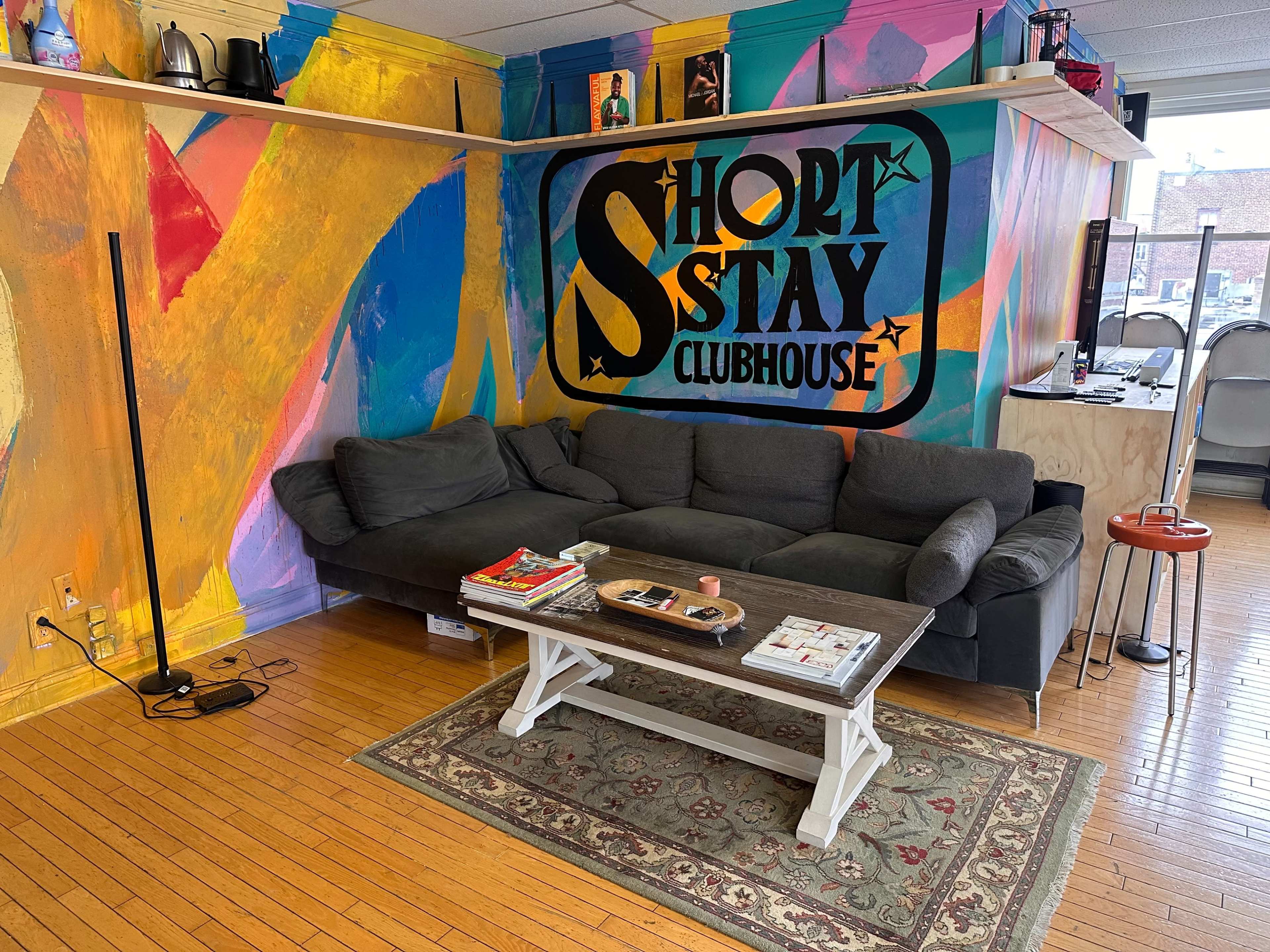 A colorful living room features a black sectional sofa, a coffee table with magazines, and a wall painted with the words "SHORT STAY CLUBHOUSE."