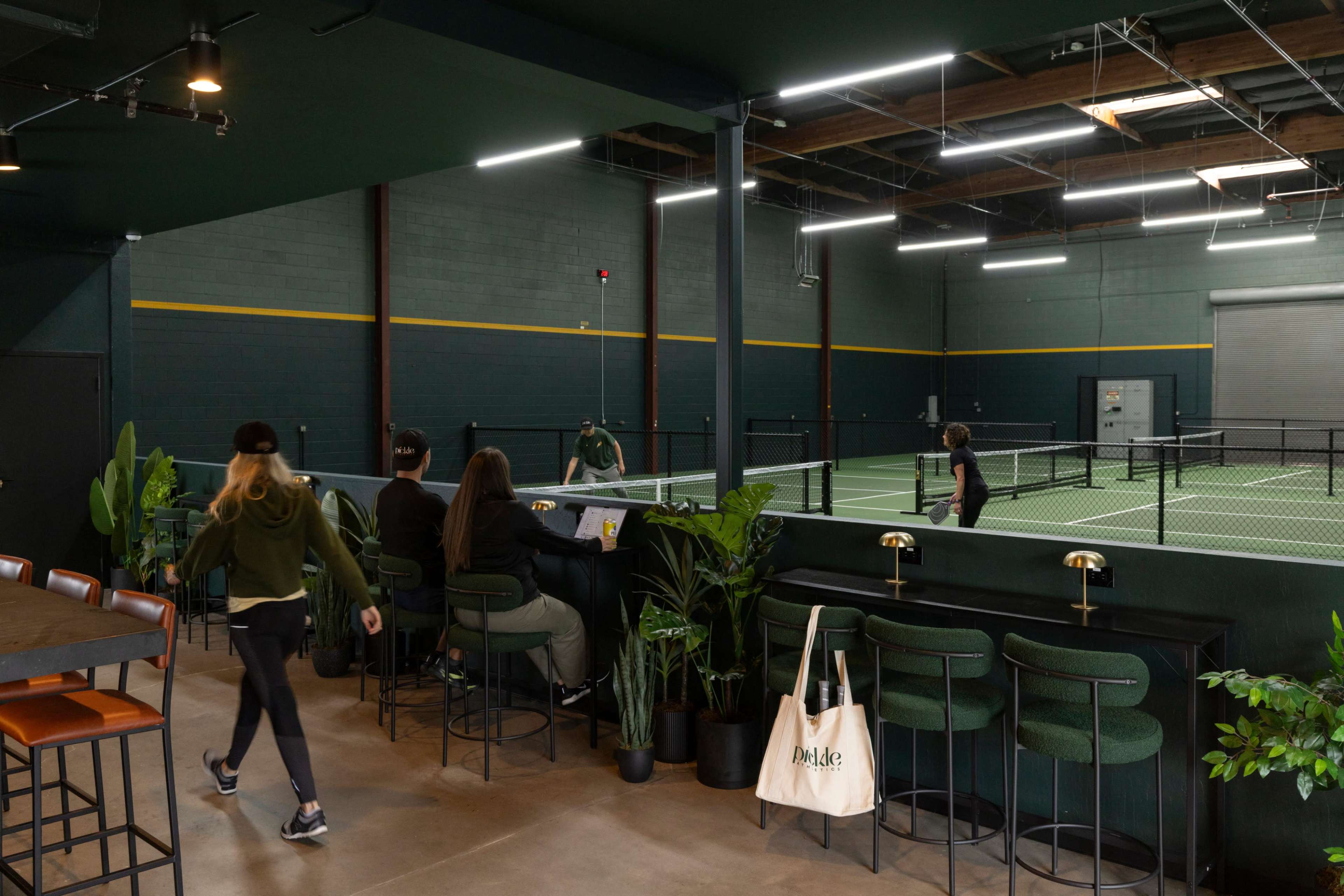 The image shows an indoor sports facility with a green and black color scheme, featuring a tennis court in the background and a bar area with patrons seated at high-top tables.