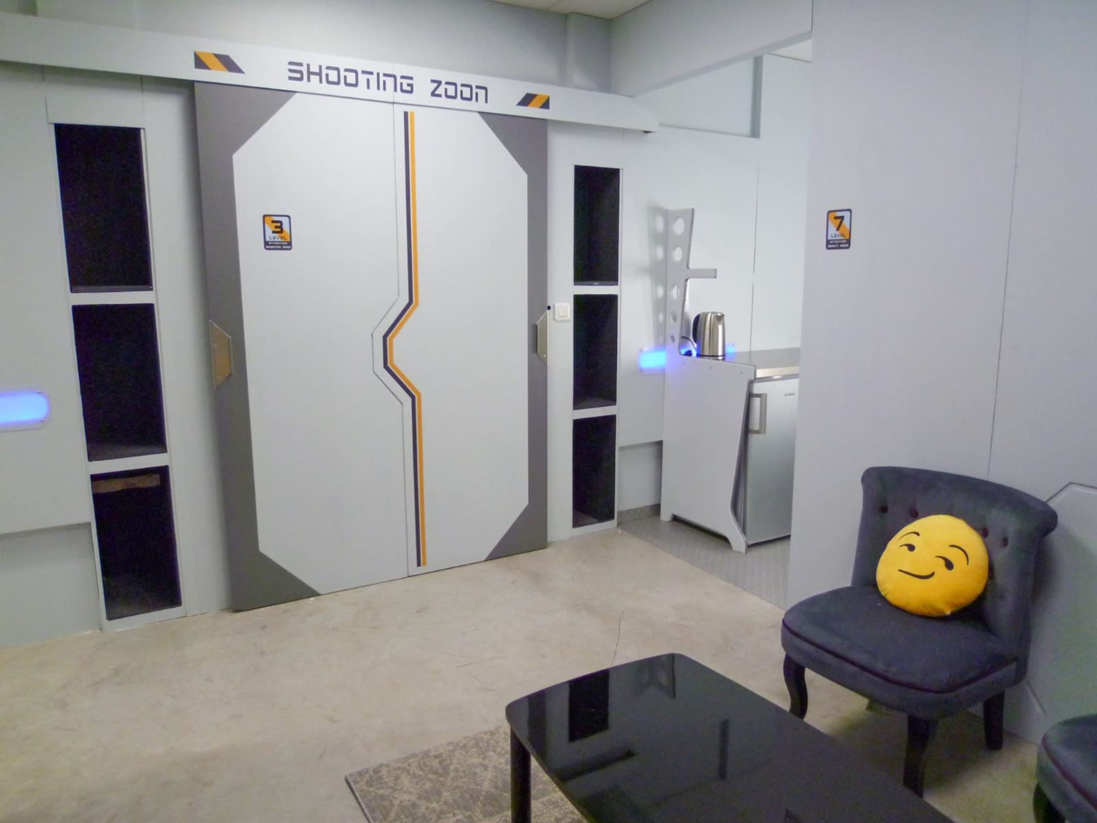 A futuristic room with a metallic door labeled "Shooting Zone," a gray upholstered chair with a smiley face pillow, and a kitchenette area in the background.