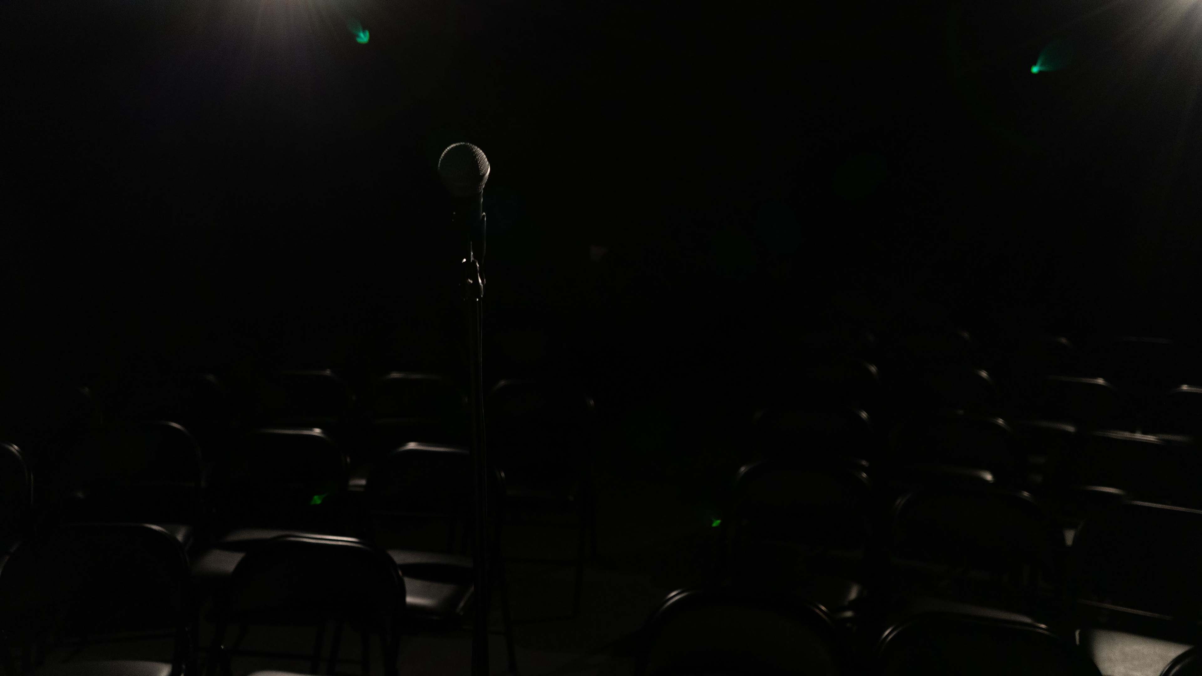 A single microphone stands on a dark stage, surrounded by empty black chairs.