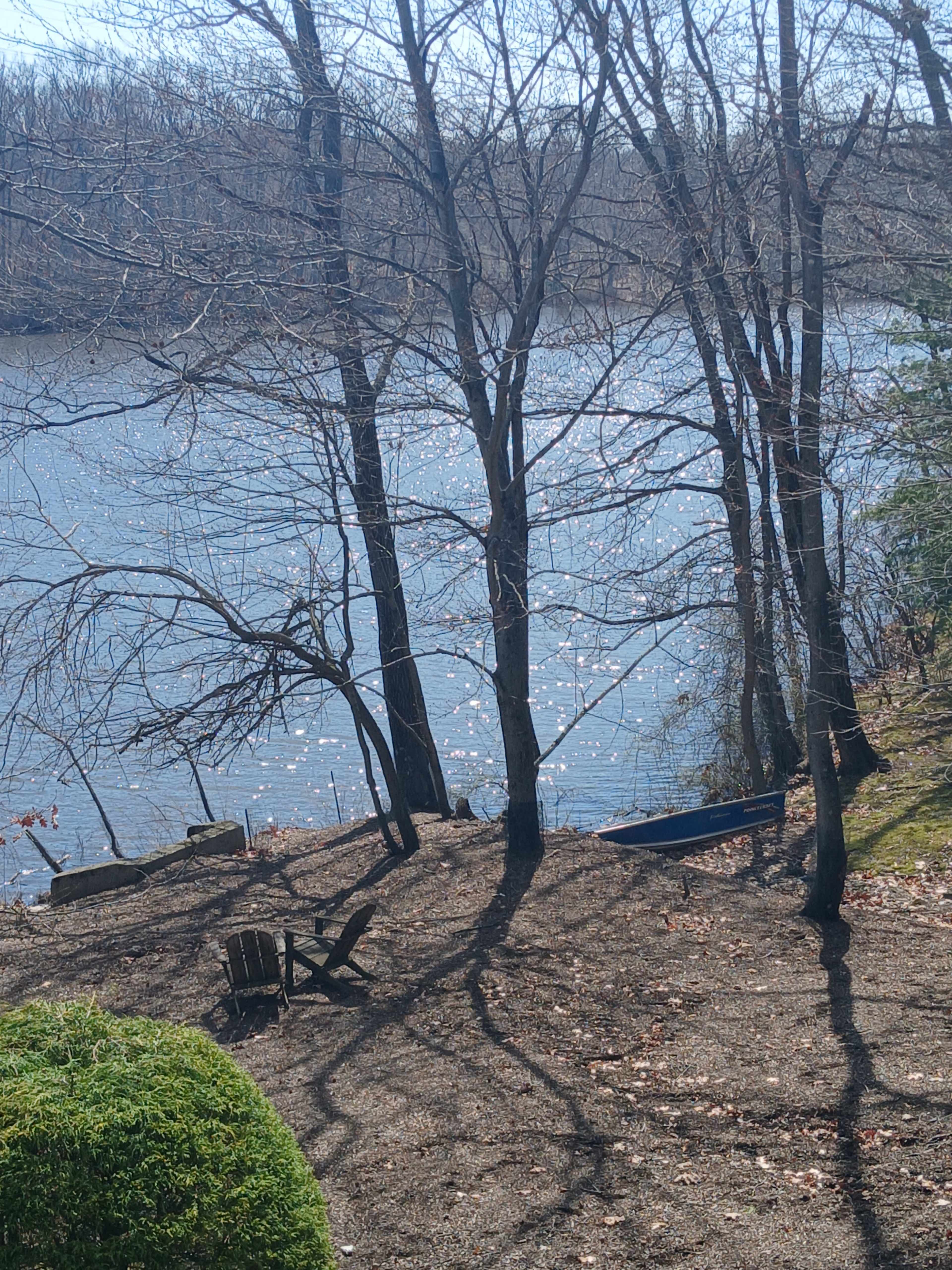 The image shows a view of a riverbank with bare trees, a wooden chair, and a small boat anchored by the water.
