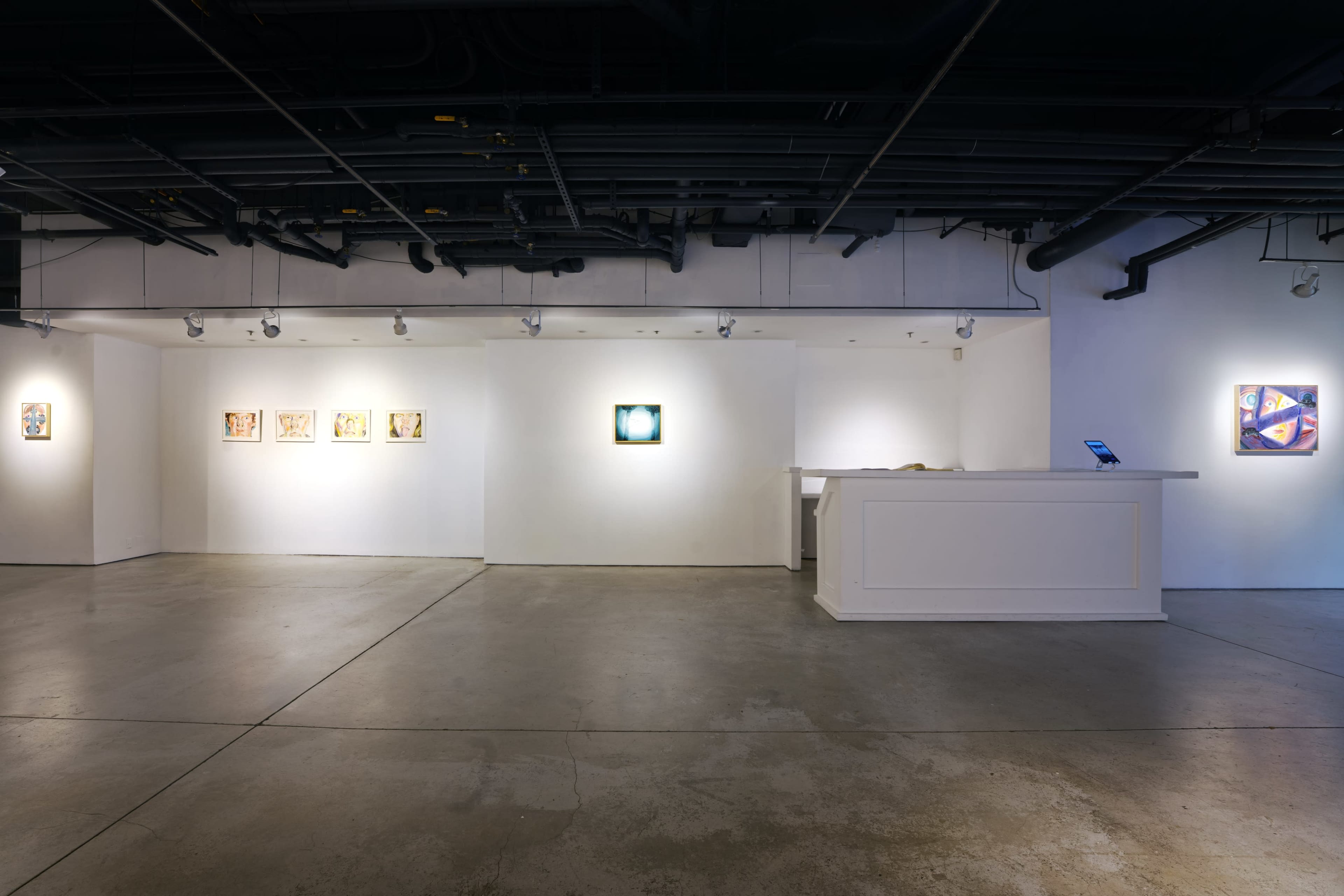 The image shows a contemporary art gallery interior with white walls, a concrete floor, and a reception desk, featuring several framed artworks displayed on the walls.