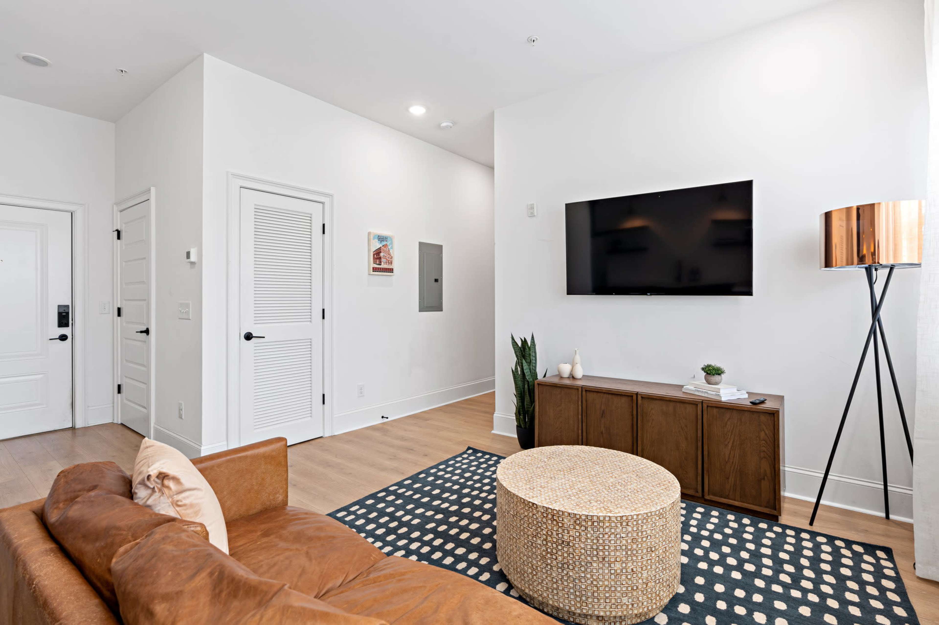 A modern living room features a leather sofa, a round woven coffee table, and a wall-mounted television.