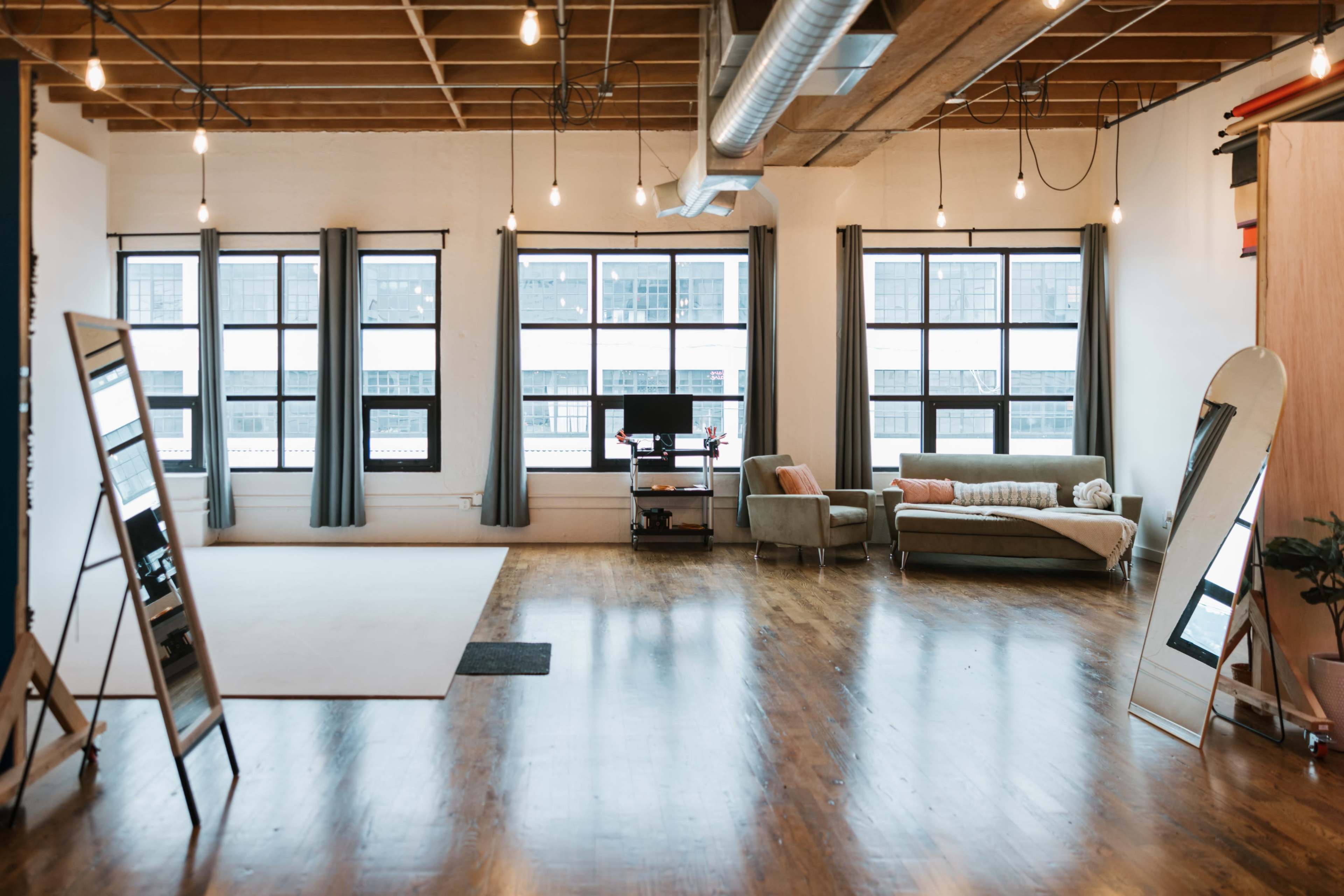 A spacious, well-lit studio with large windows, wooden floors, a green couch, and a mirror set up on an easel.