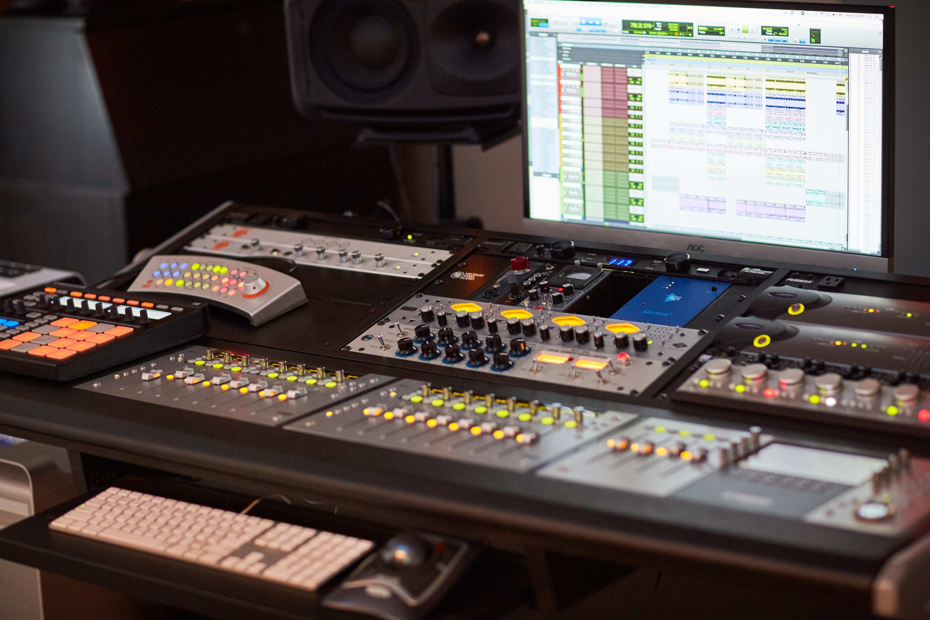 The image shows a music production workstation featuring a large control surface, digital audio interface, and a computer monitor displaying audio editing software.