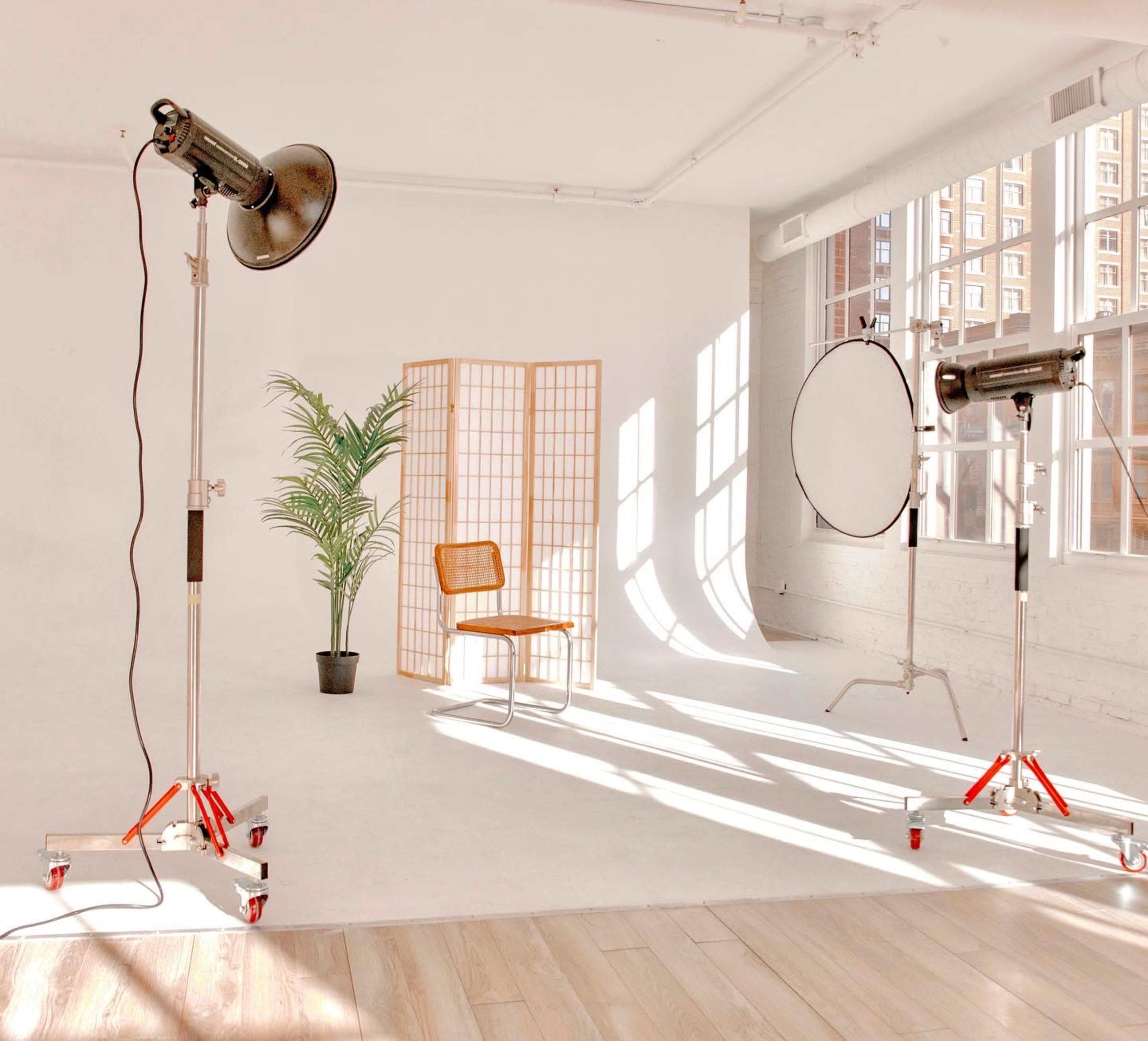 A well-lit photography studio featuring a wooden chair, a potted plant, a folding screen, and two studio lights beside a reflective disc.