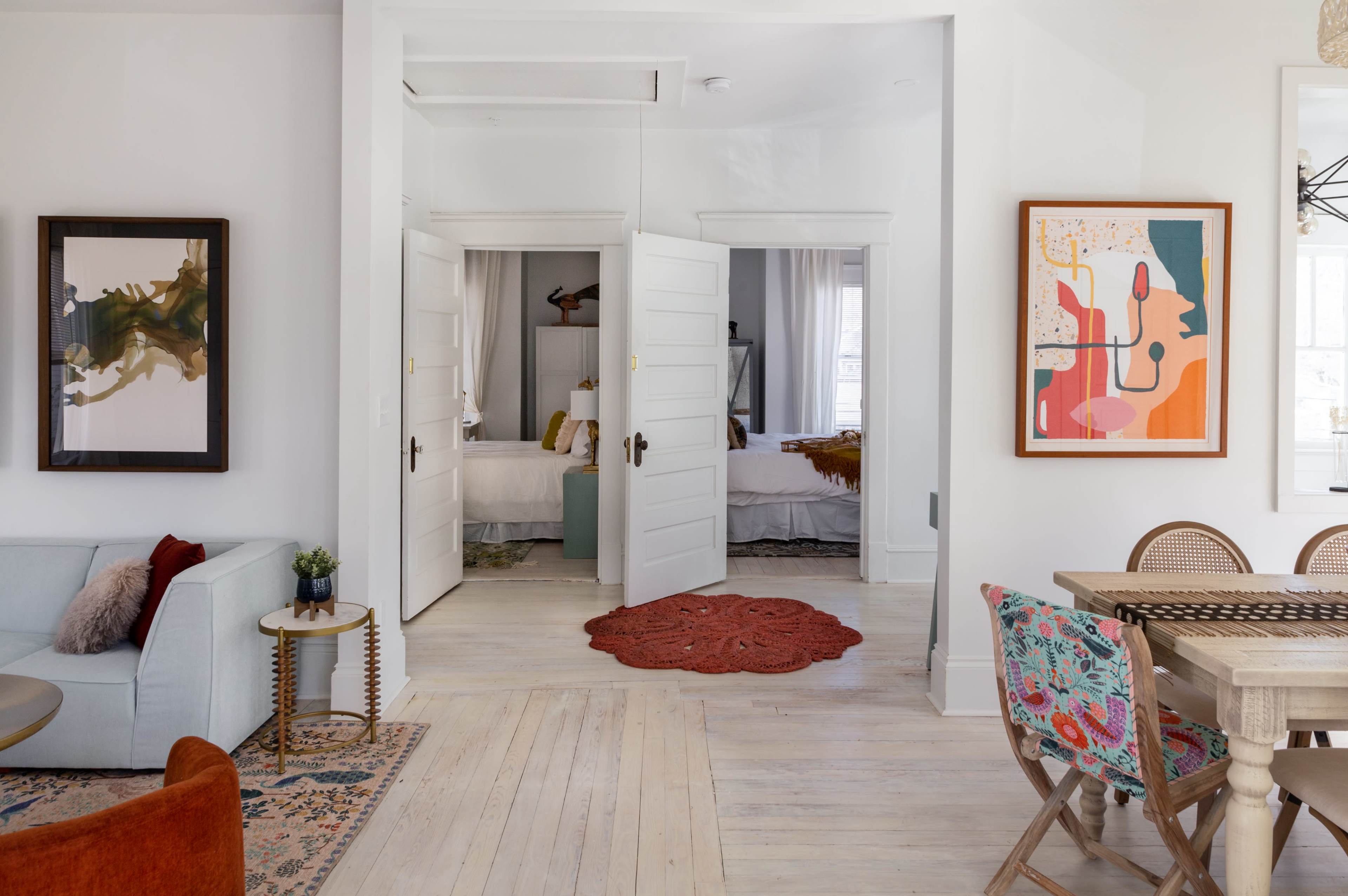 A bright, open living space features a light-colored couch, a doorway with double doors leading to another room, and colorful artwork on the walls.