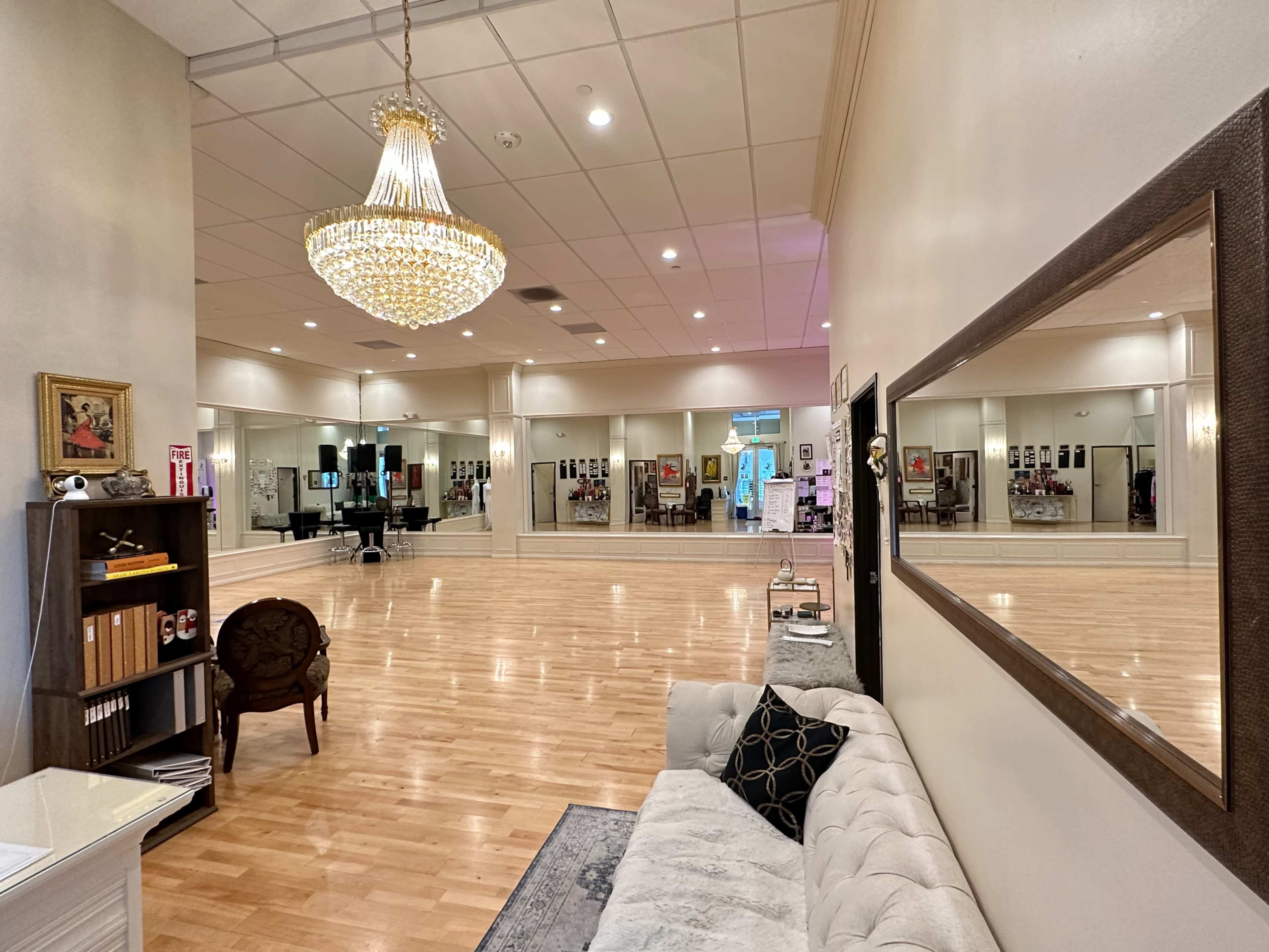 A spacious dance studio featuring a wooden floor, a chandelier, mirrors on the walls, and a seating area with a couch.