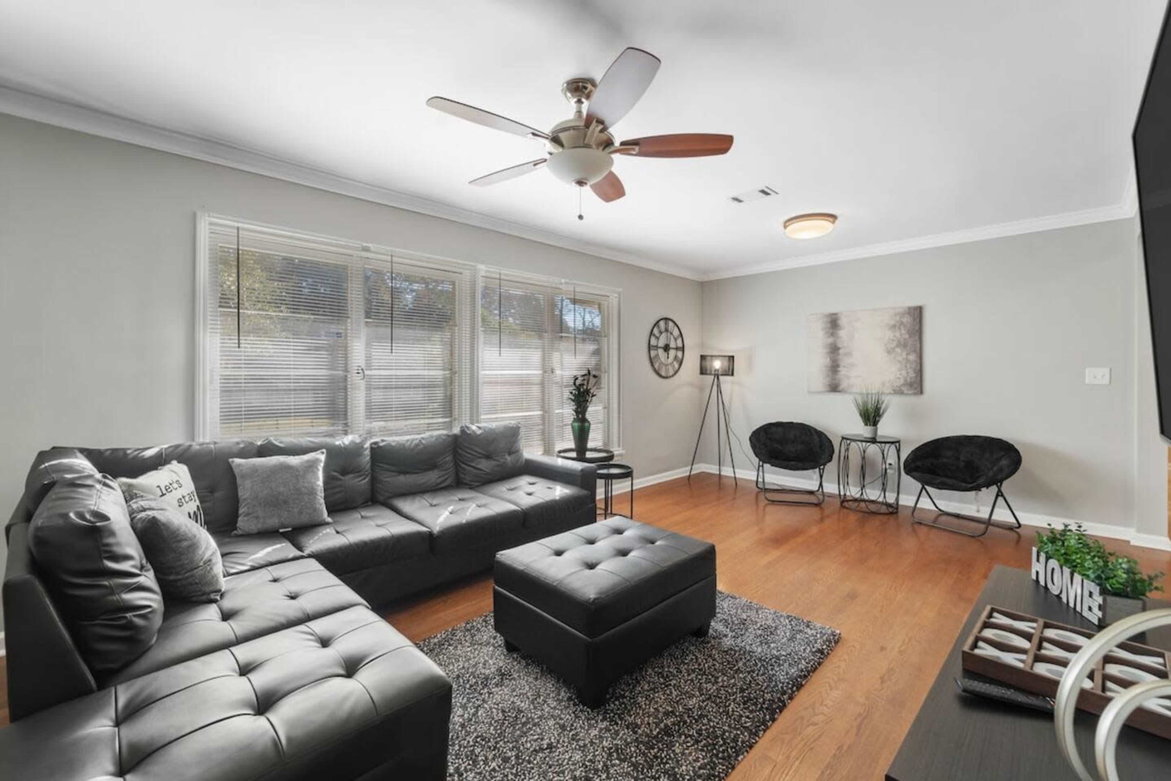 The living room features a sectional sofa, a coffee table, and two accent chairs arranged around a rug, with large windows allowing natural light to enter.