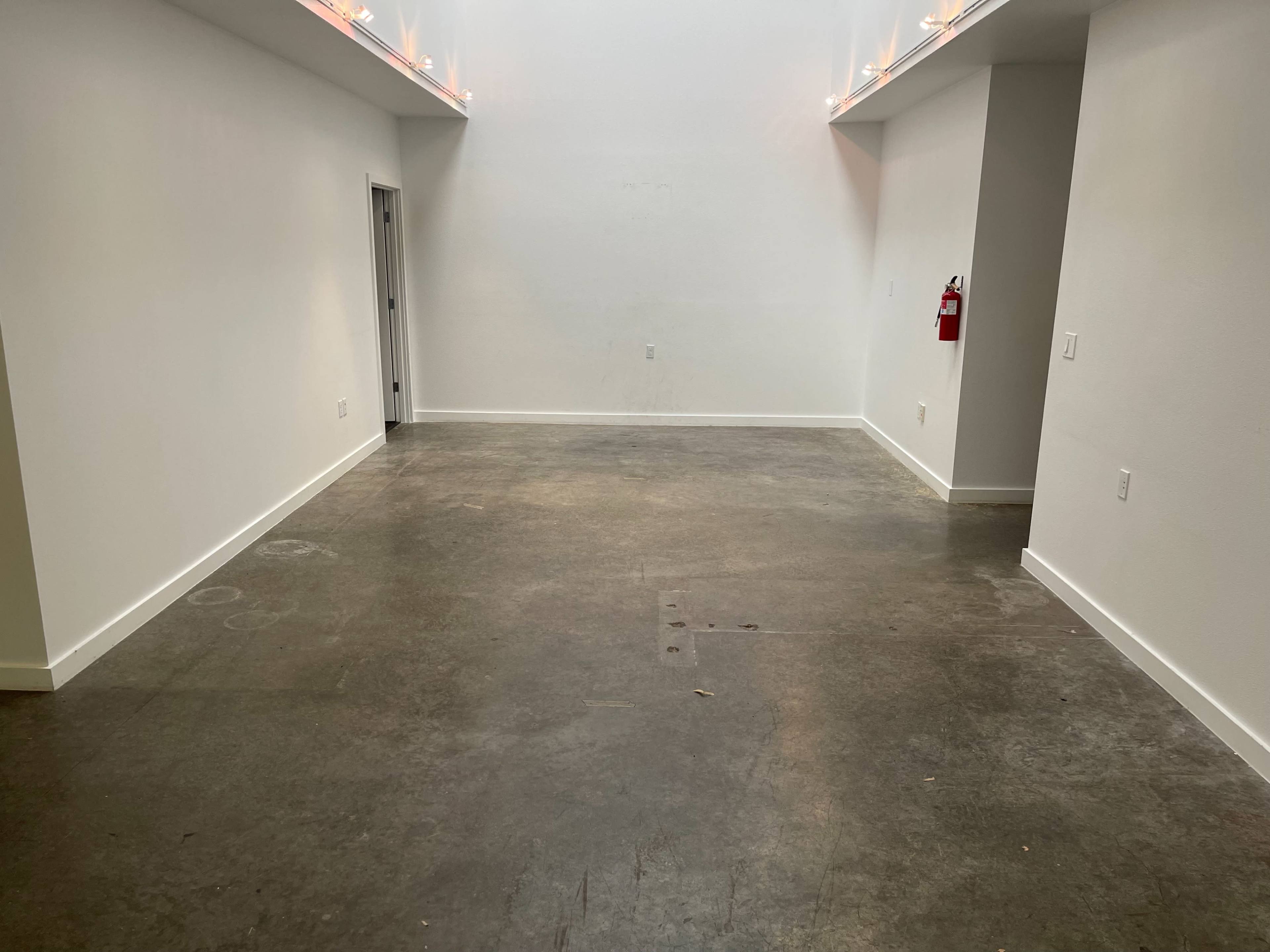 The image shows a minimalistic interior space with a concrete floor, white walls, and a single fire extinguisher on the right side.