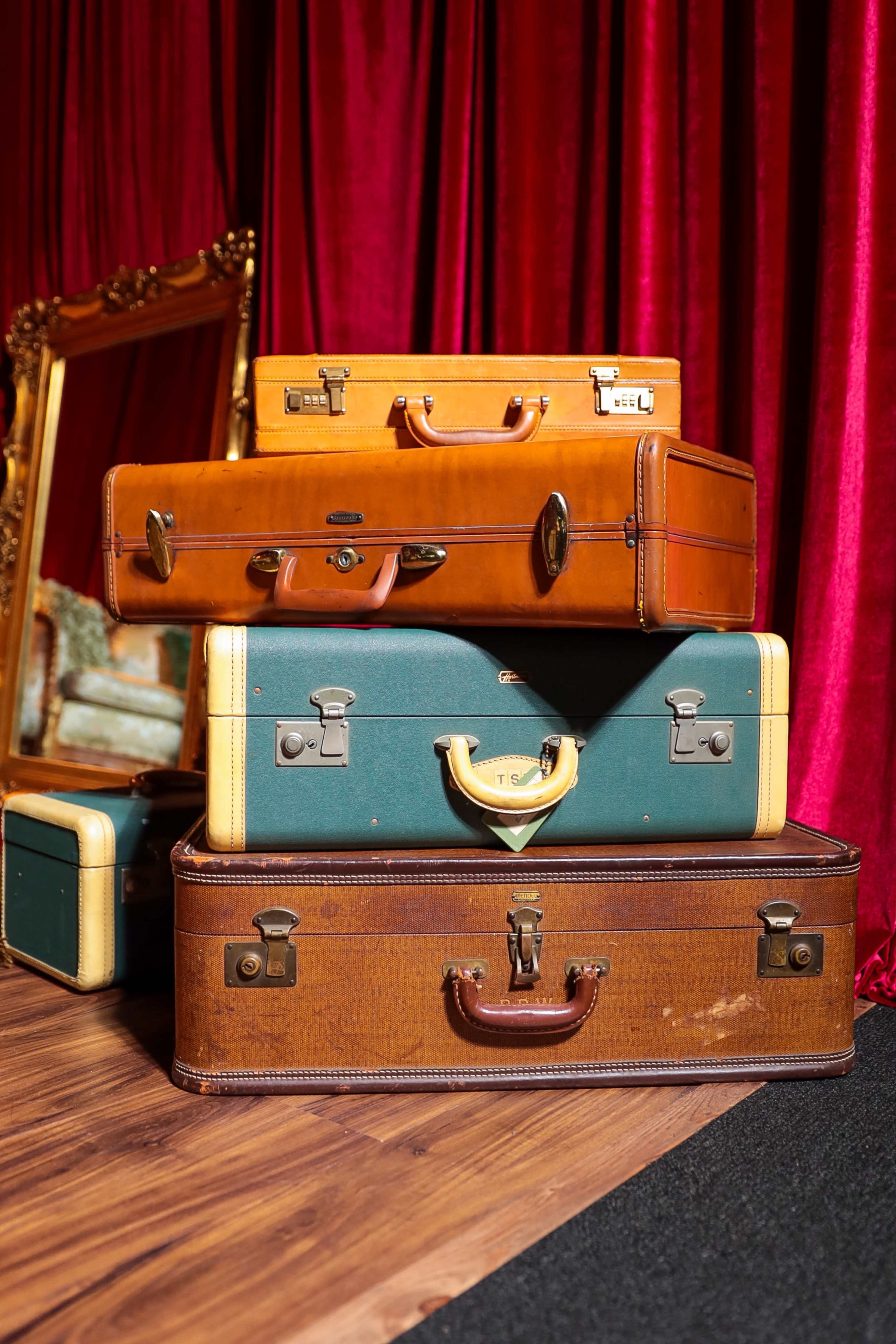A stack of vintage suitcases in various colors is arranged next to a large ornate mirror against a red curtain backdrop.