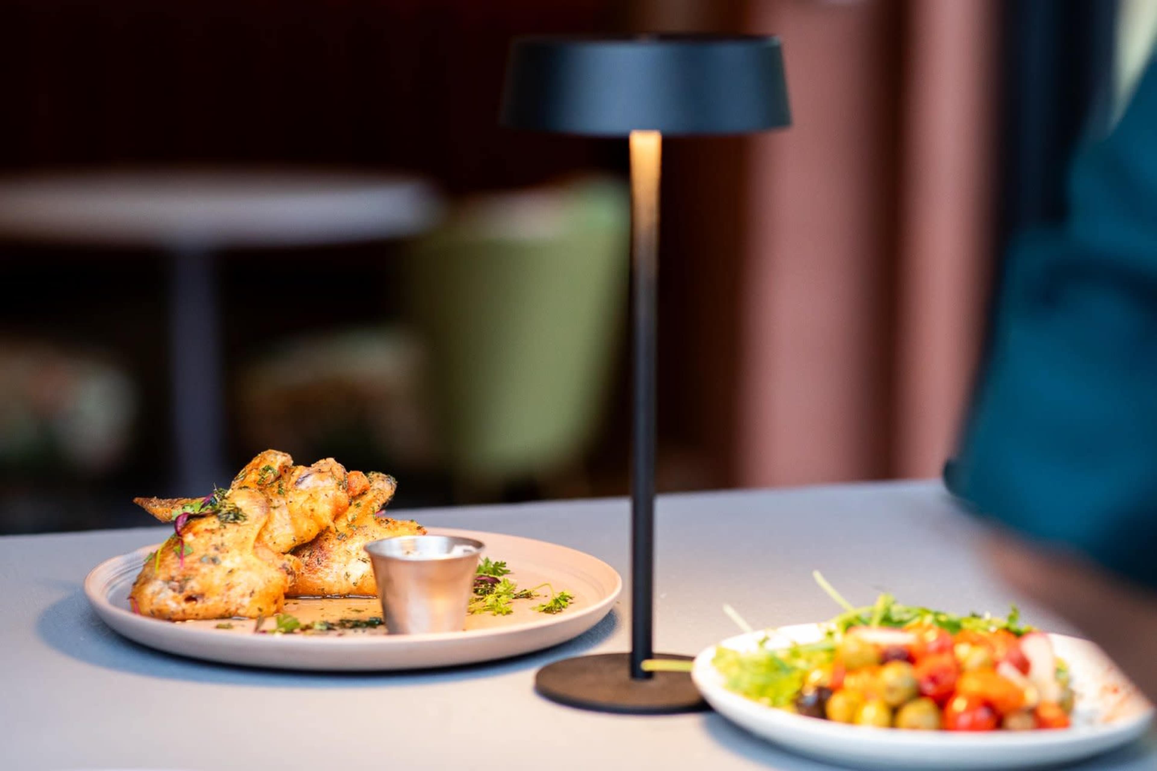 A plate of chicken wings with a dipping sauce is placed next to a salad of mixed vegetables on a table with a lamp.