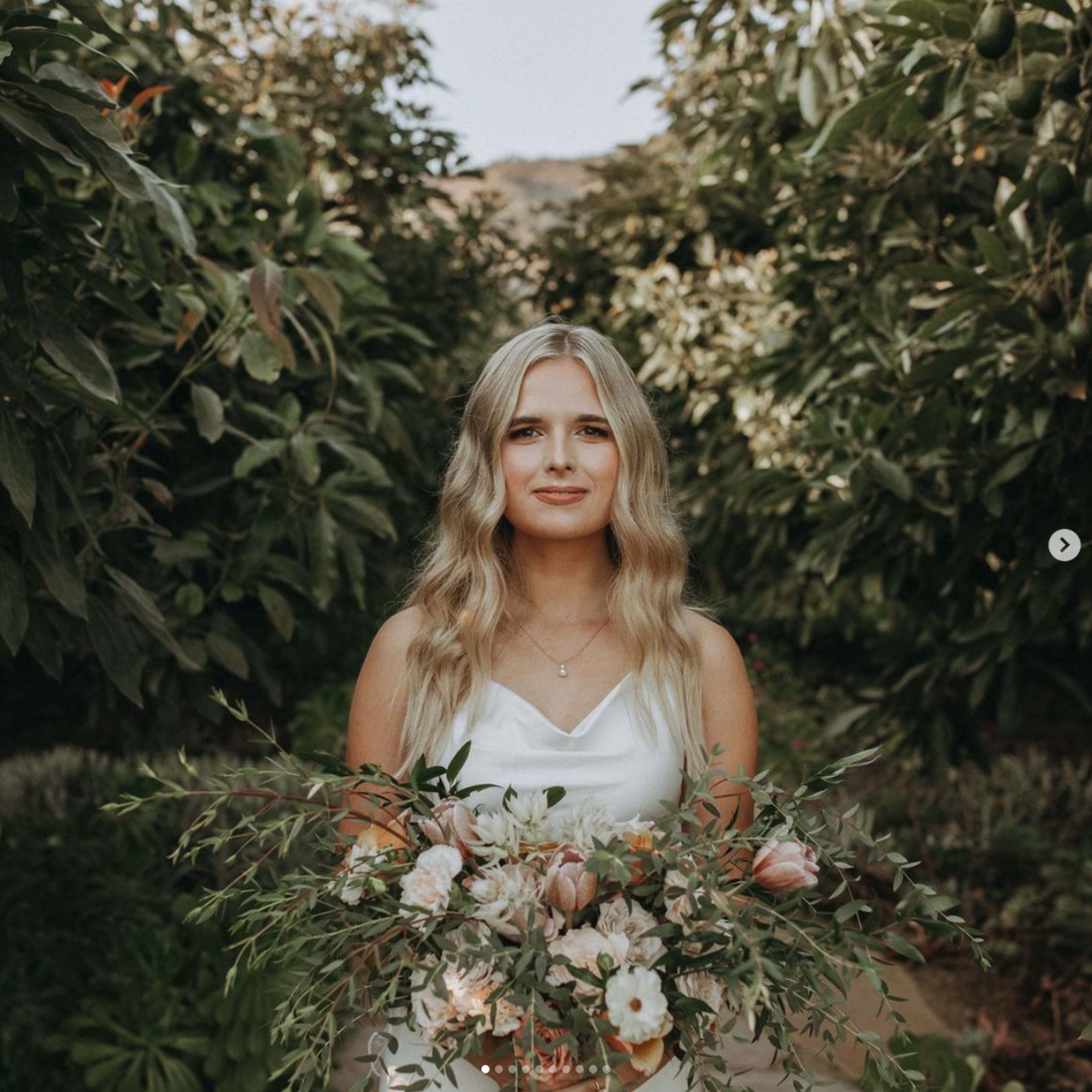 A woman holds a bouquet of flowers while standing in a garden surrounded by greenery.