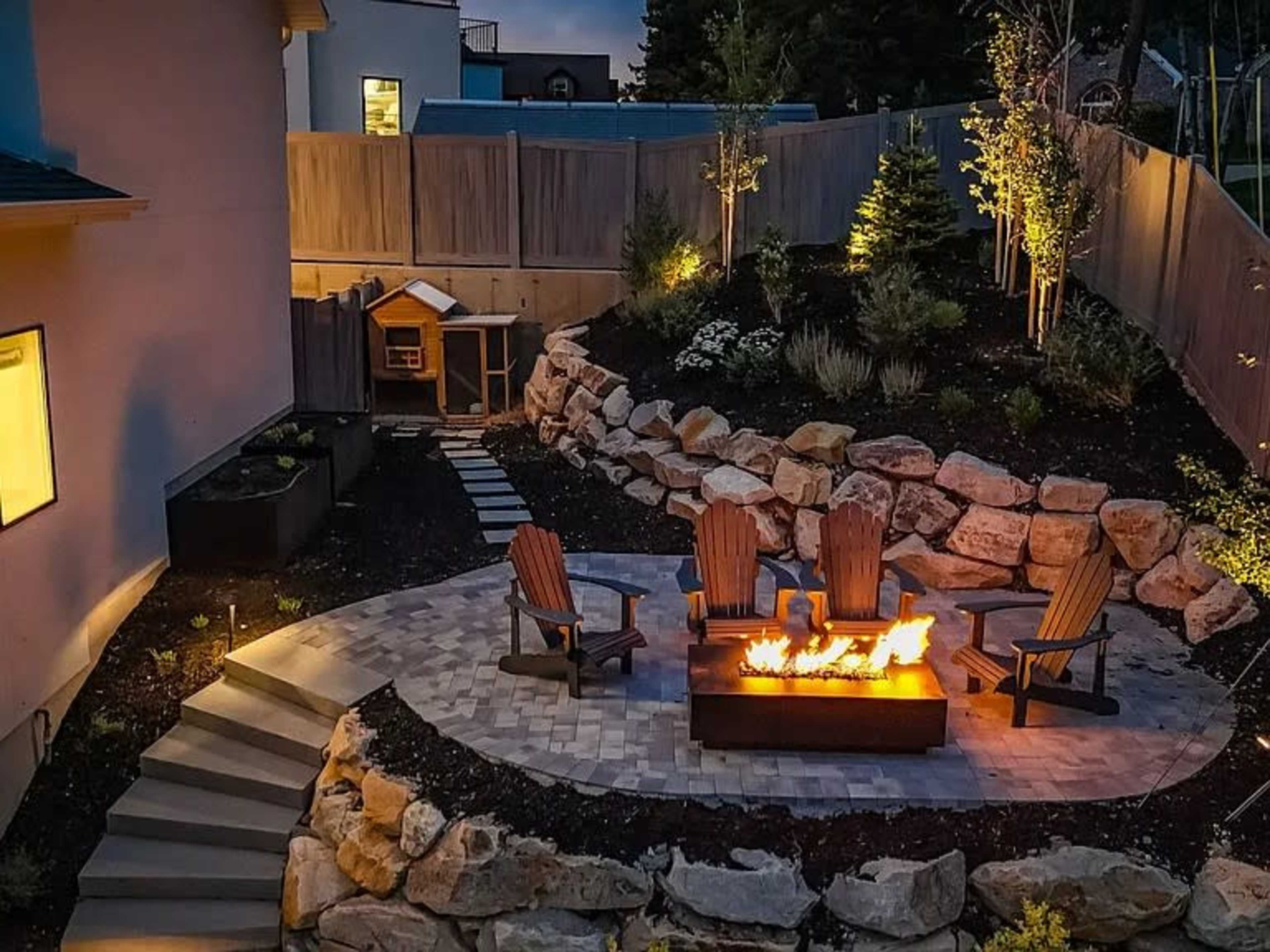 The image shows a backyard patio featuring a fire pit surrounded by four wooden chairs, stone landscaping, and a small shed in the background.