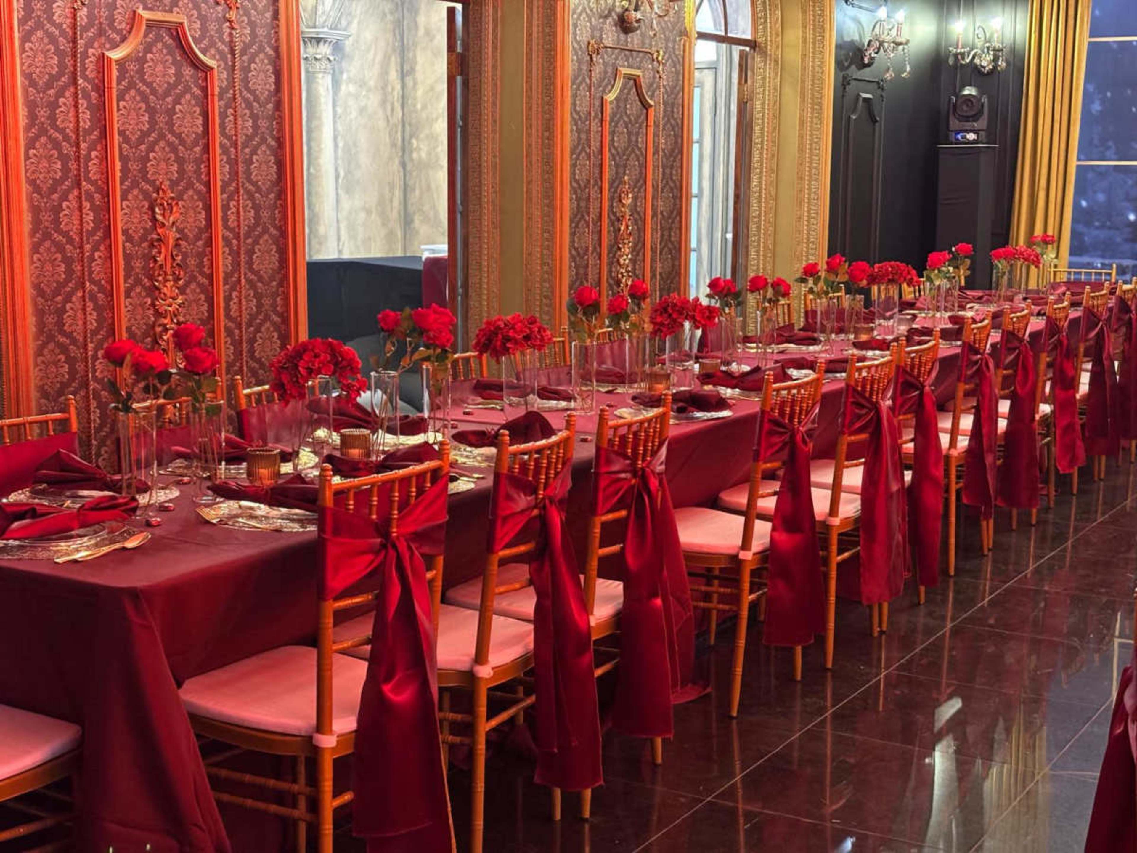 A long banquet table is elegantly set with red tablecloths, adorned with glass vases of red roses, and surrounded by gold chairs tied with red sashes.