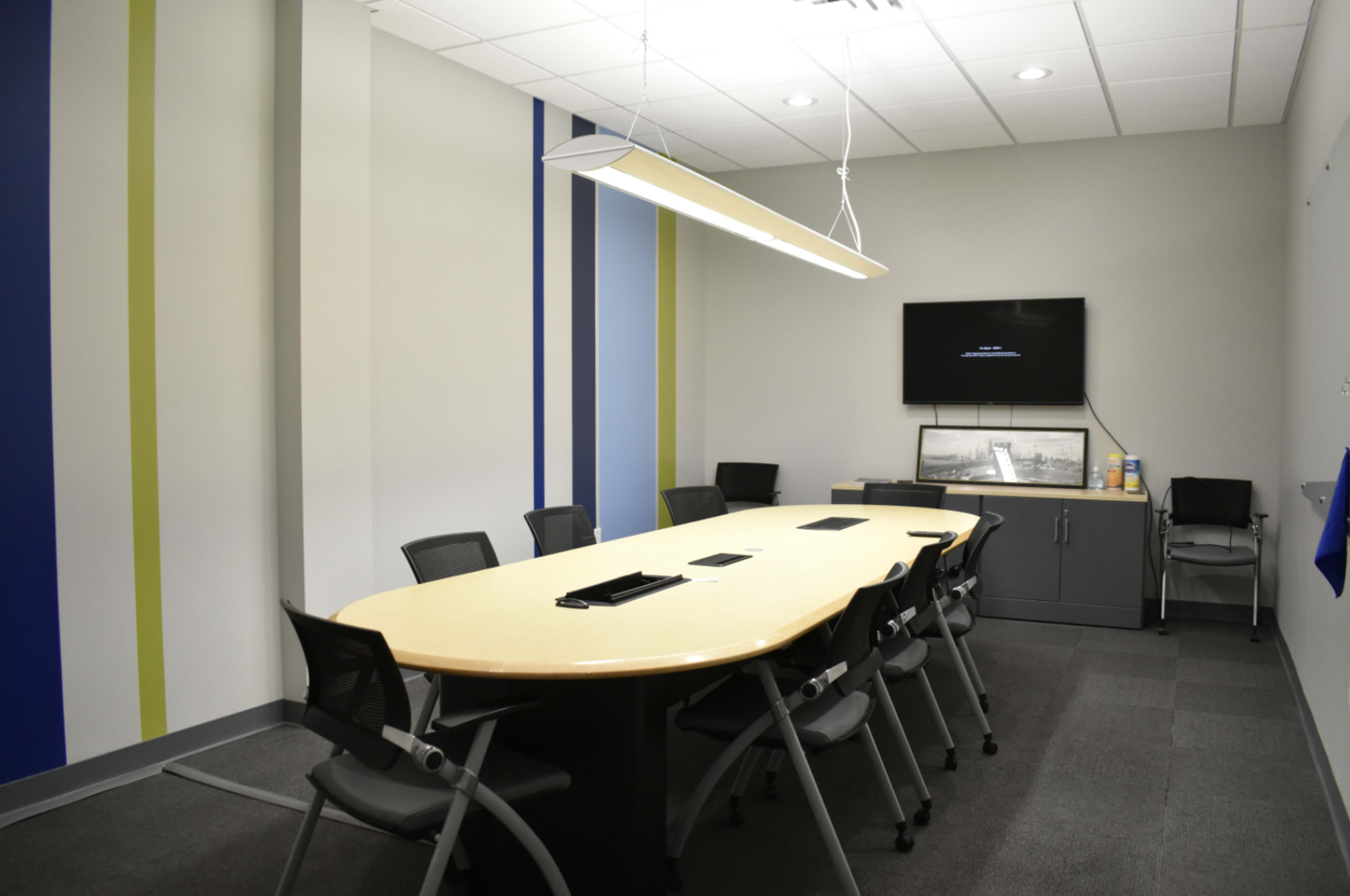 A modern conference room features a large oval table surrounded by black chairs, with a television mounted on the wall and colored stripes decorating the walls.