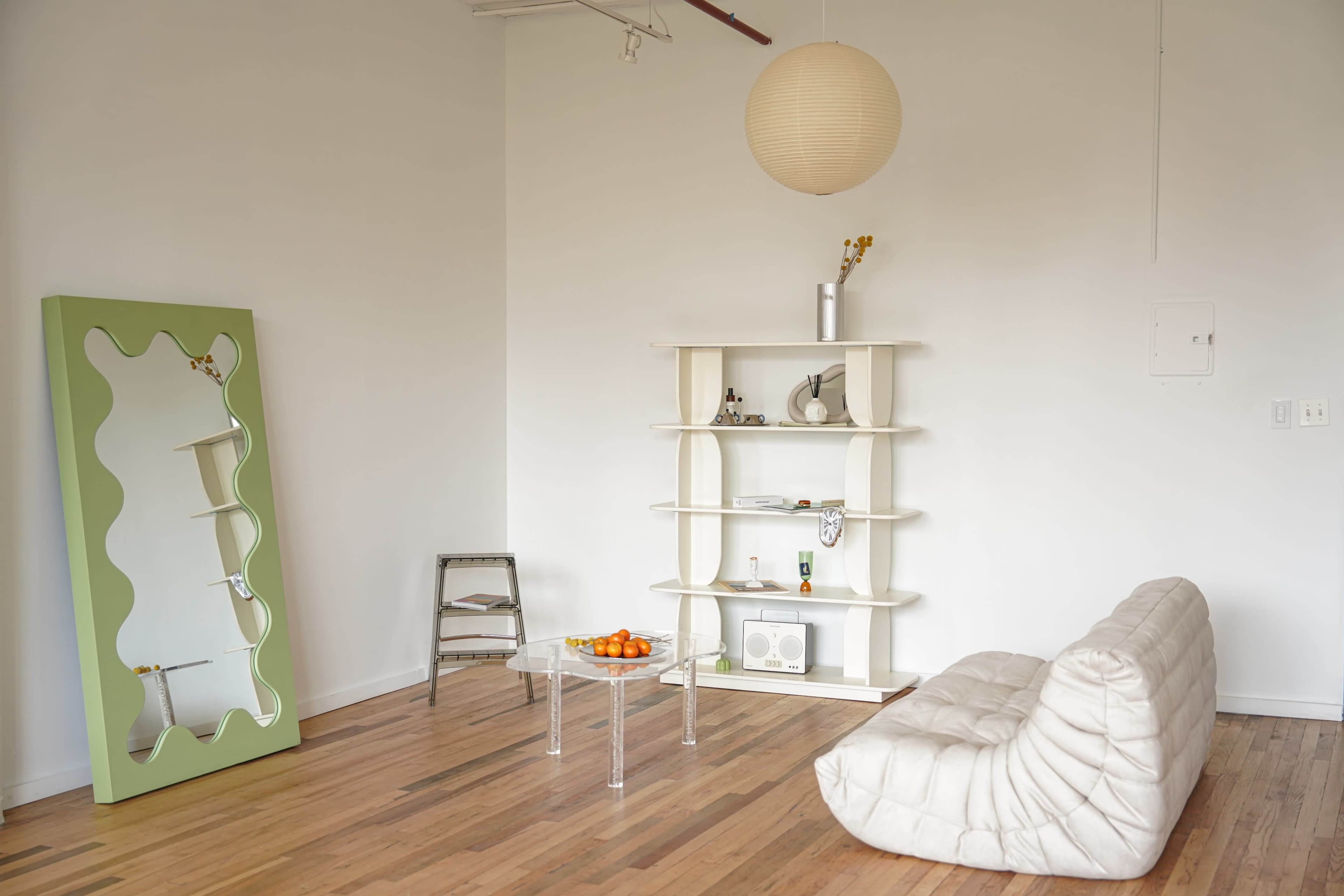 A minimalist room features a white tufted sofa, a green mirror, a shelf with decorative items, and a bowl of oranges on a clear table.