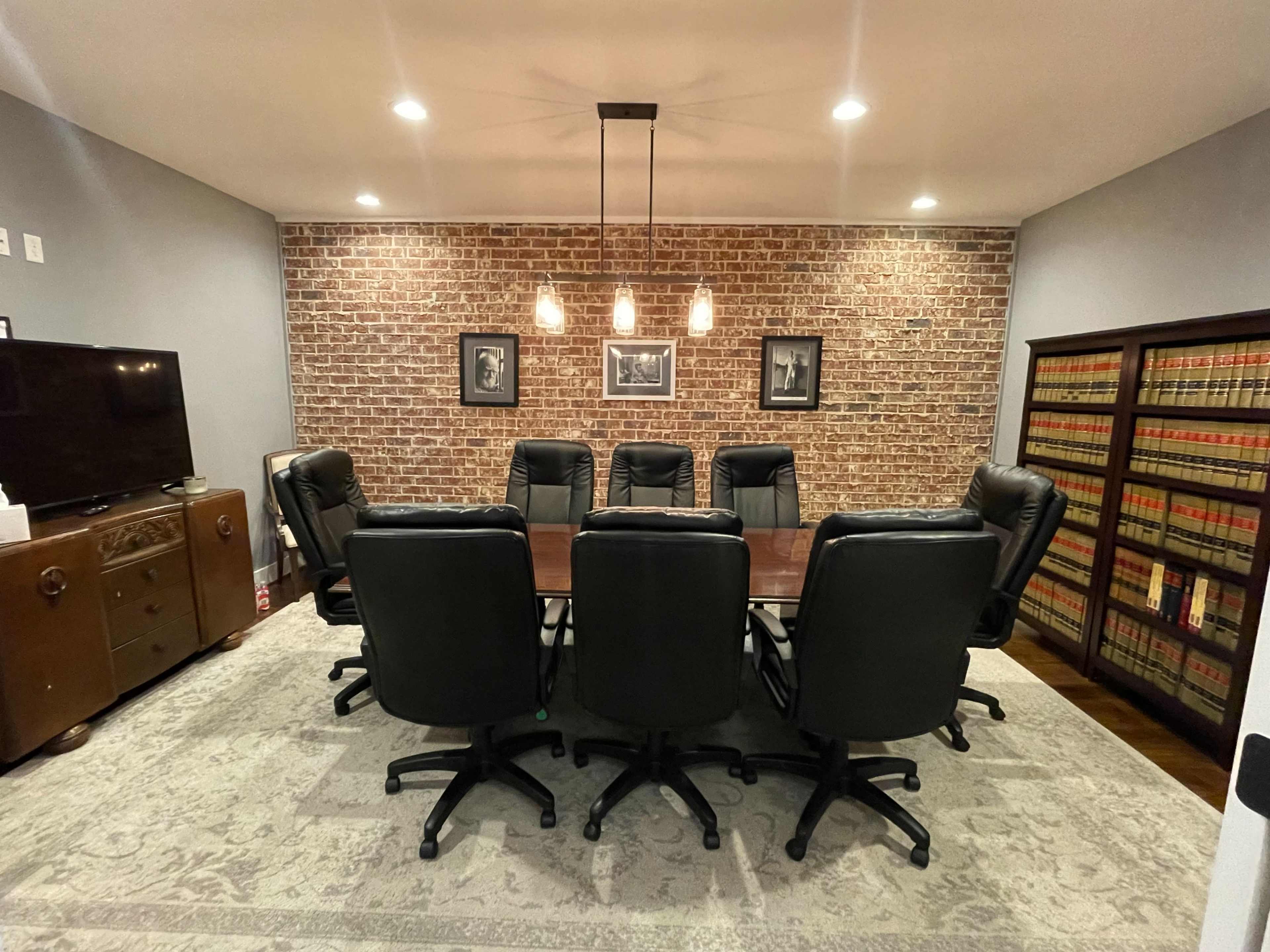 A conference room featuring a large rectangular table surrounded by eight black leather chairs, against a brick wall with framed pictures.