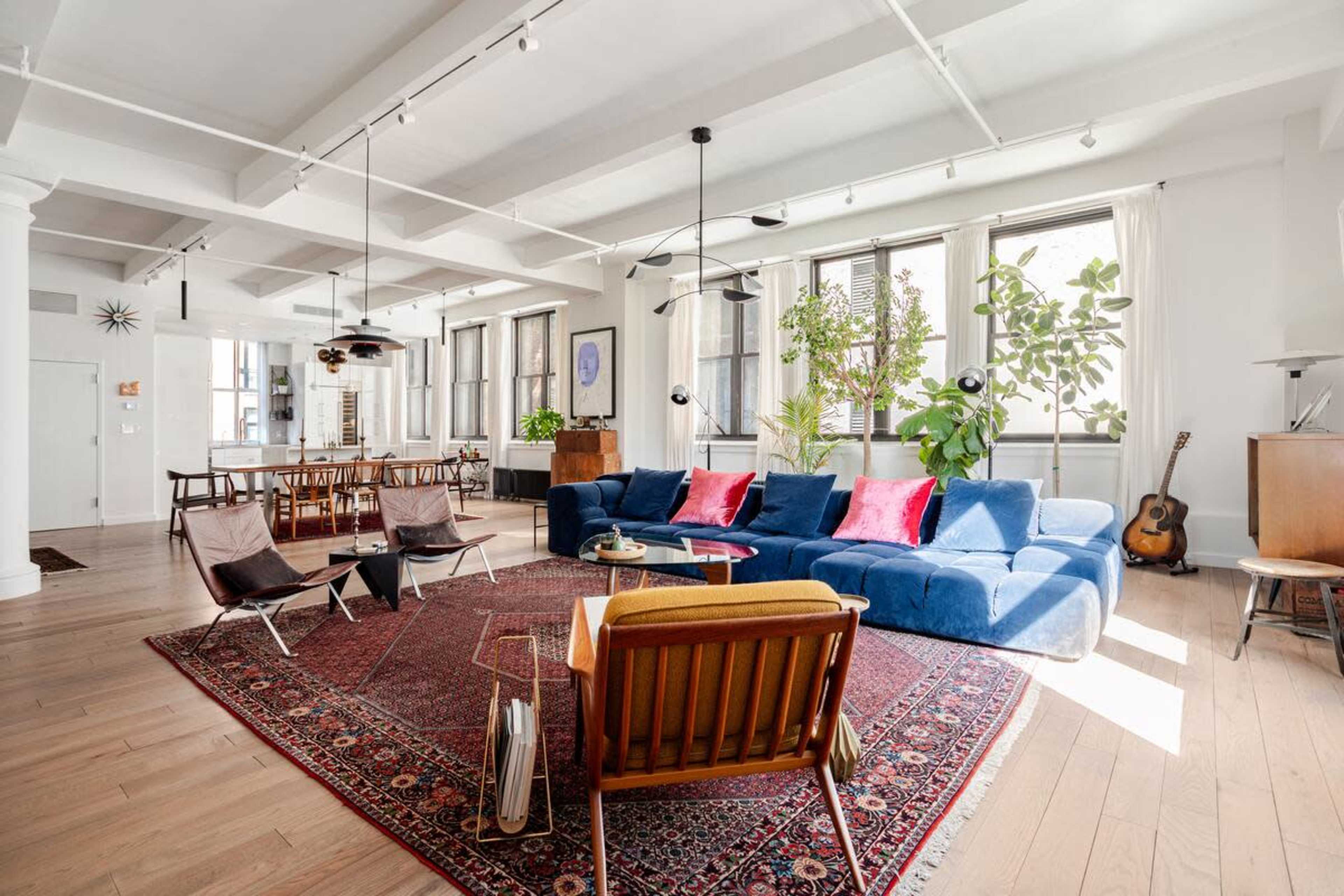 The image shows a spacious, modern living area with large windows, a blue sectional sofa, and a wooden dining table surrounded by chairs.