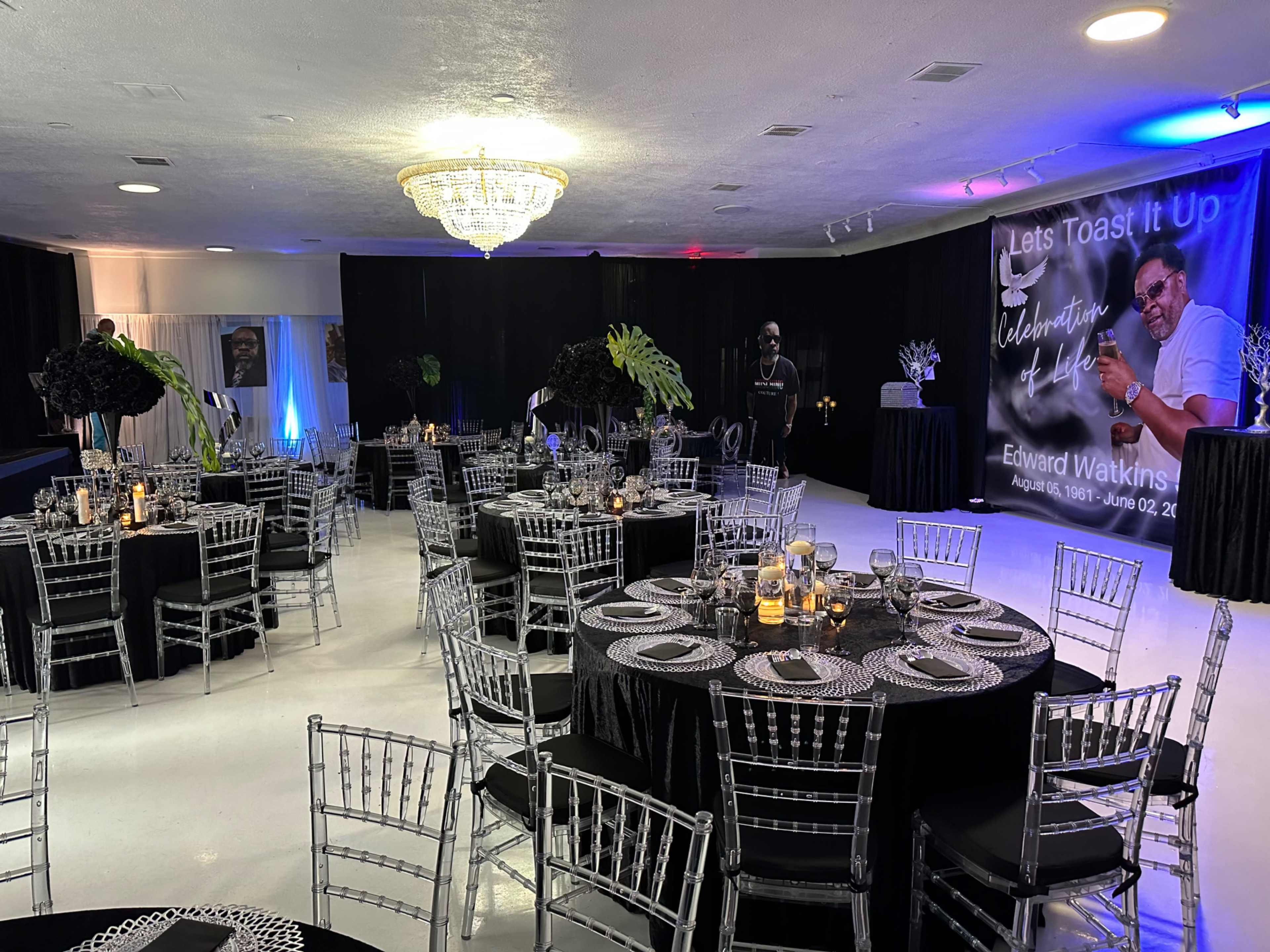 A banquet hall set up with round tables covered in black tablecloths, silver chiavari chairs, and decorative centerpieces, featuring a backdrop celebrating Edward Watkins with a large portrait.