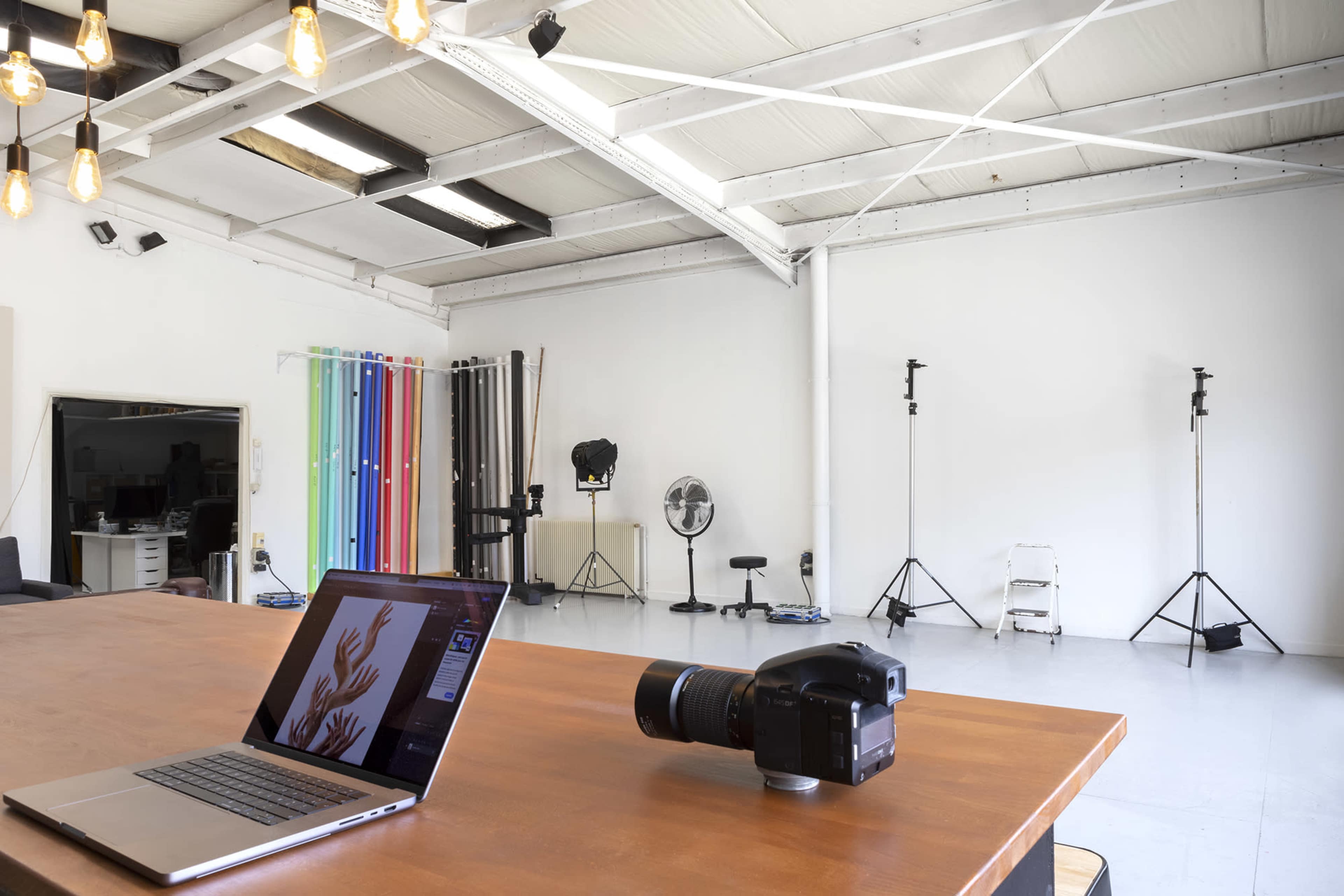 A spacious photography studio features a wooden table with a laptop and camera, surrounded by studio lighting equipment and backdrops.