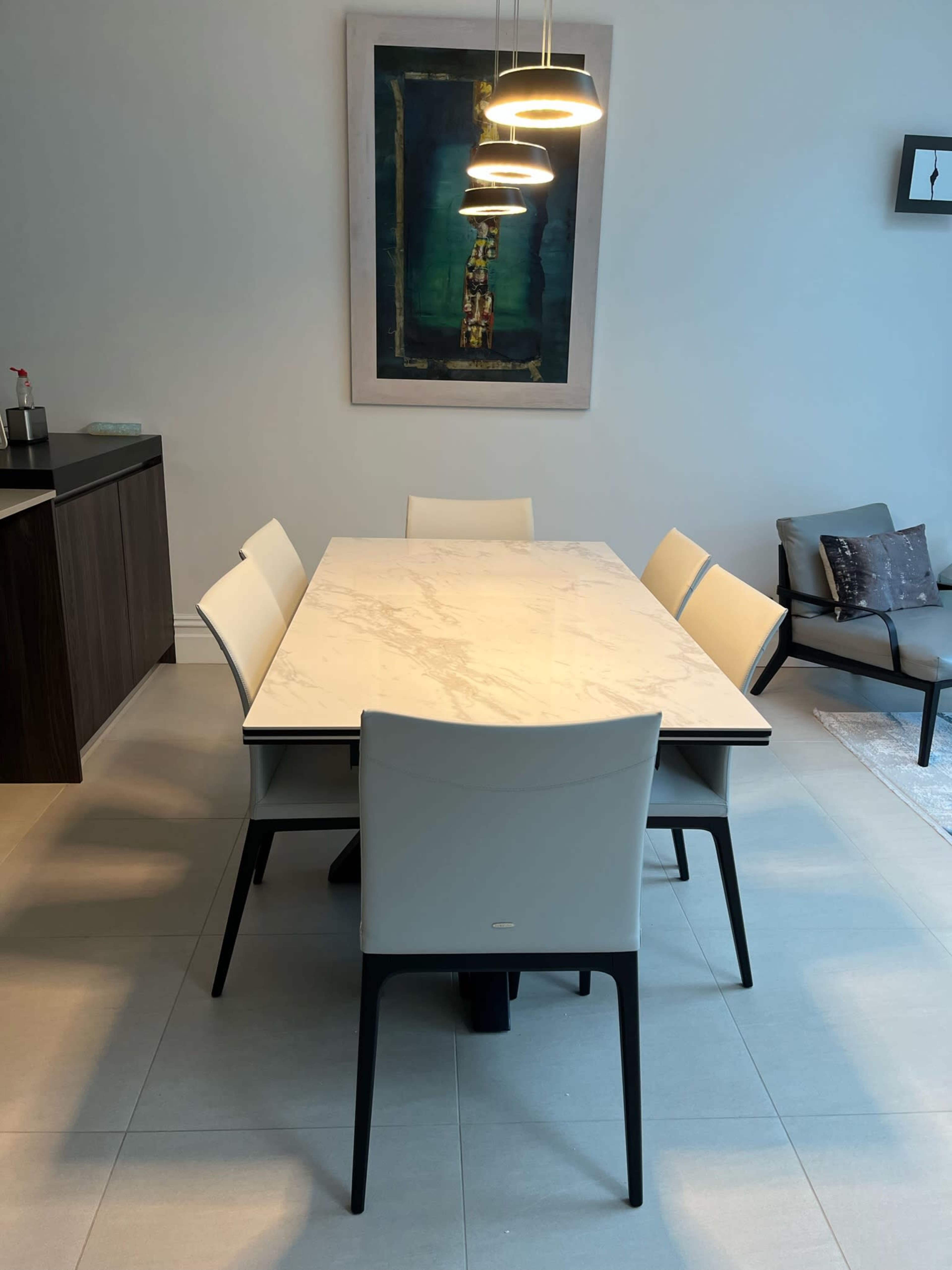 A modern dining table with six chairs is positioned under pendant lights in a minimalist dining area featuring a large piece of abstract art on the wall.