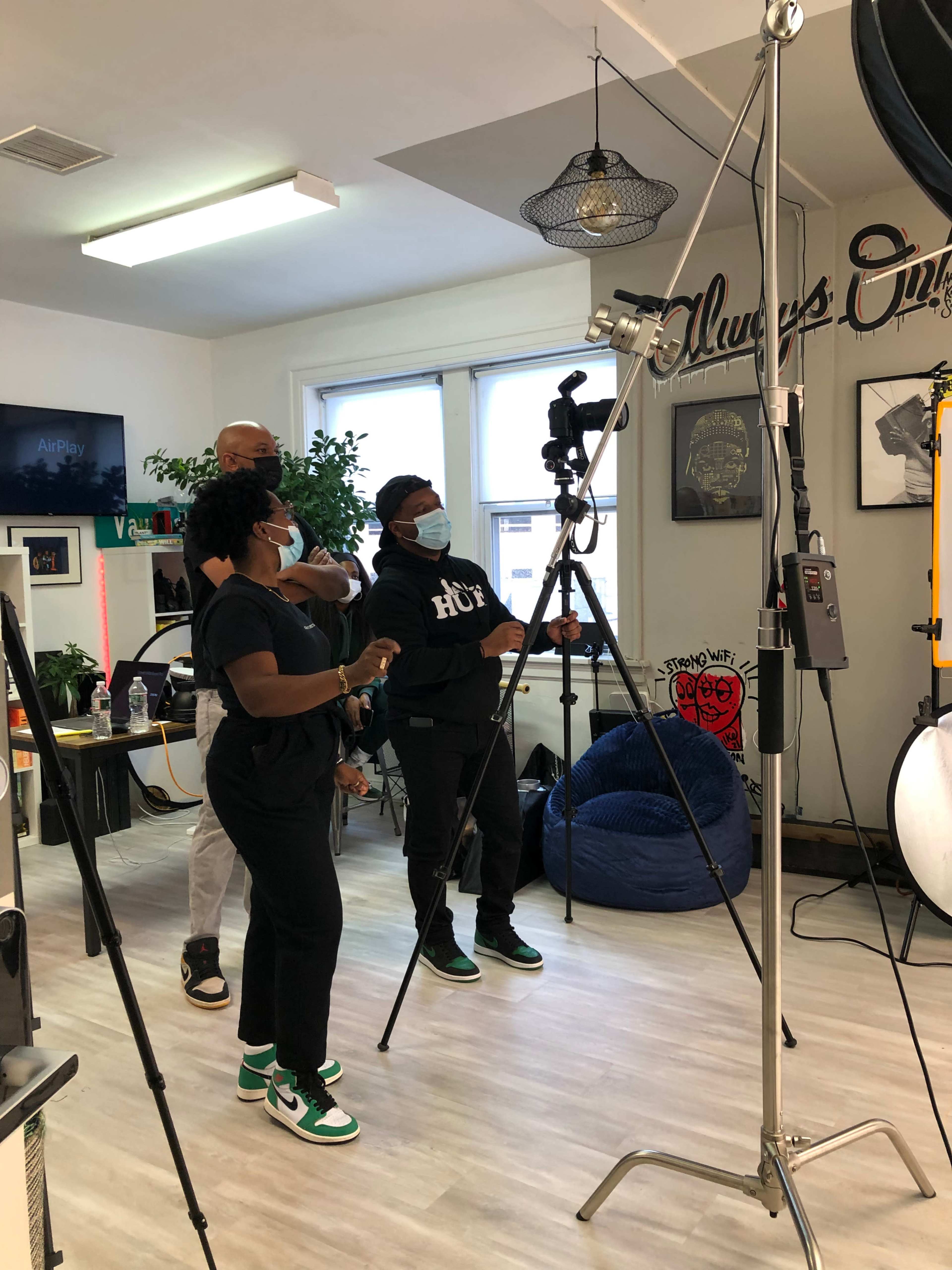 A group of three people is adjusting equipment in a studio setting, with a tripod and lighting setup in the foreground.