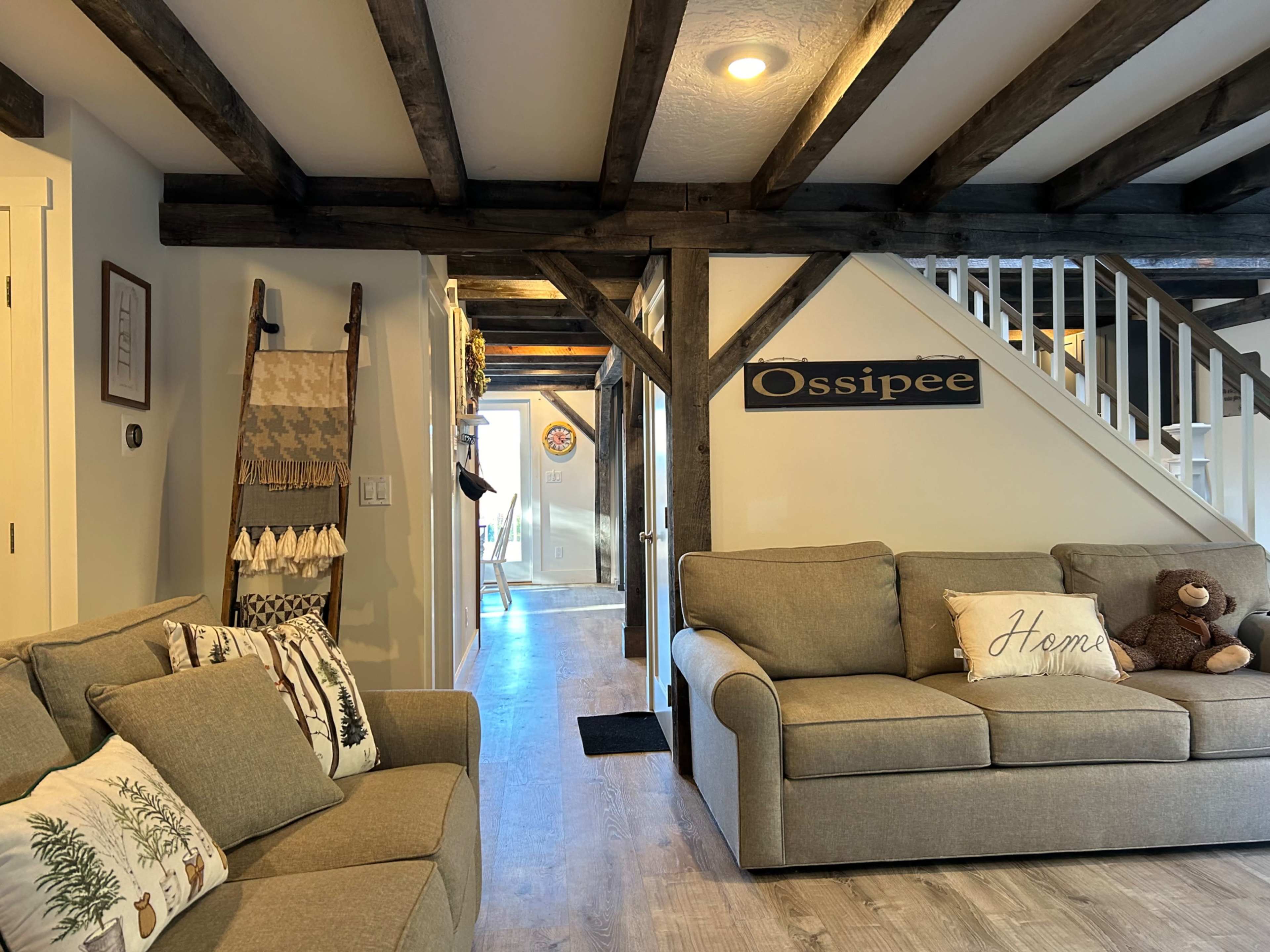 The image shows a cozy living room with a gray sofa, wood beams on the ceiling, and a staircase leading to another level.