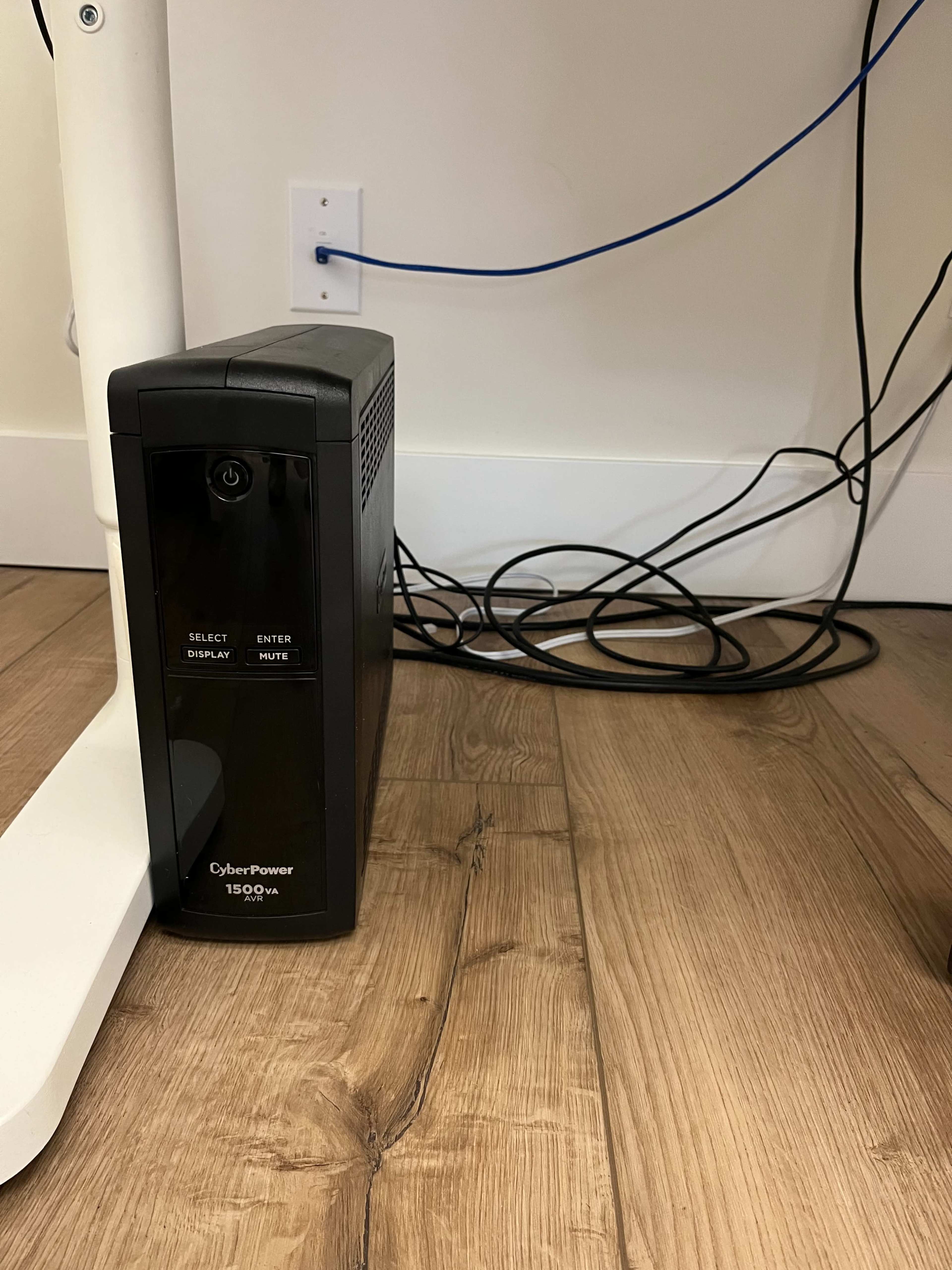 A CyberPower UPS is positioned on the floor next to a white desk leg, with various electrical cables scattered around it.