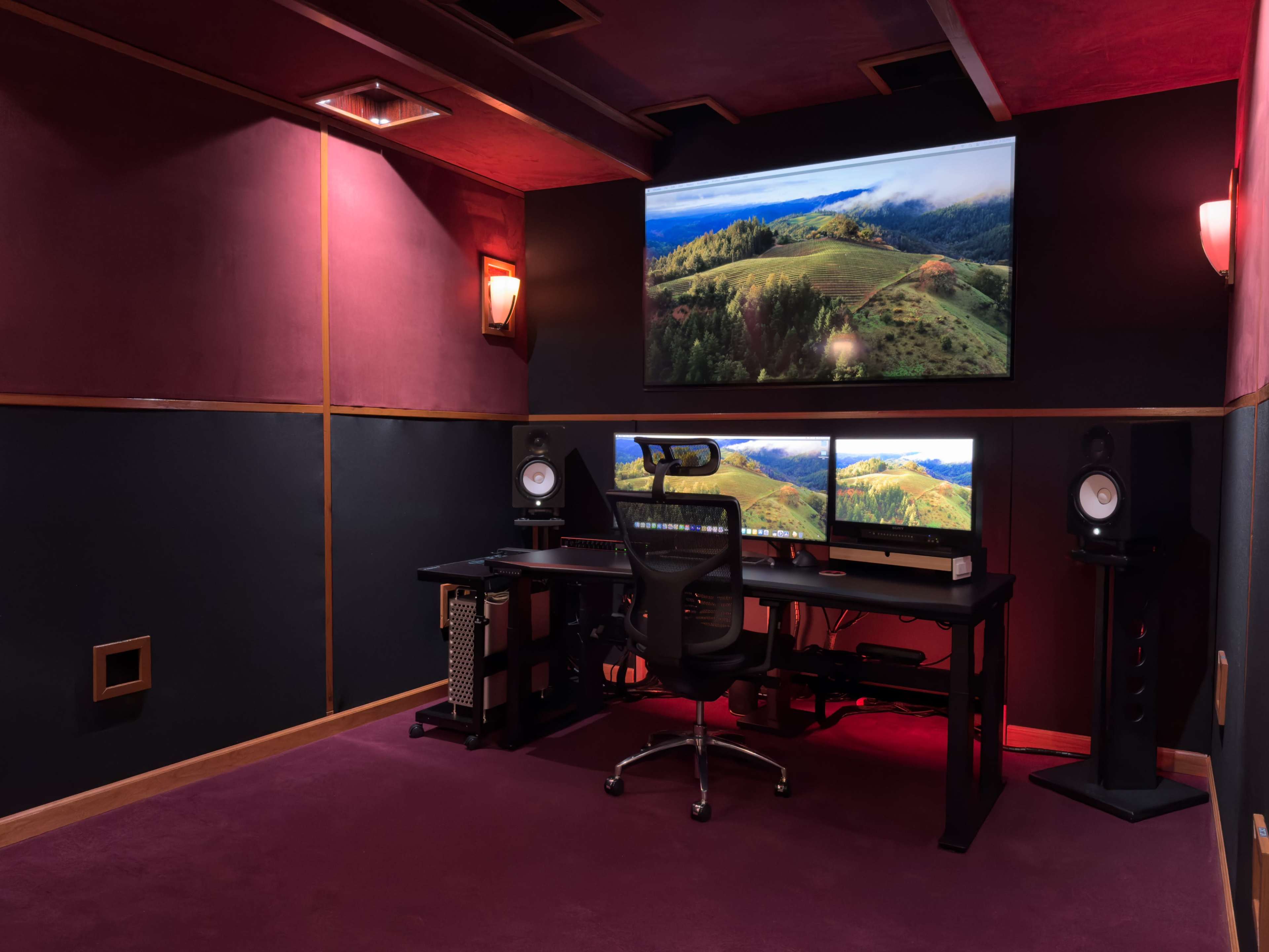 A sound studio featuring a desk with two computer monitors, a chair, and large speakers, all situated against dark walls with a large screen displaying a landscape.