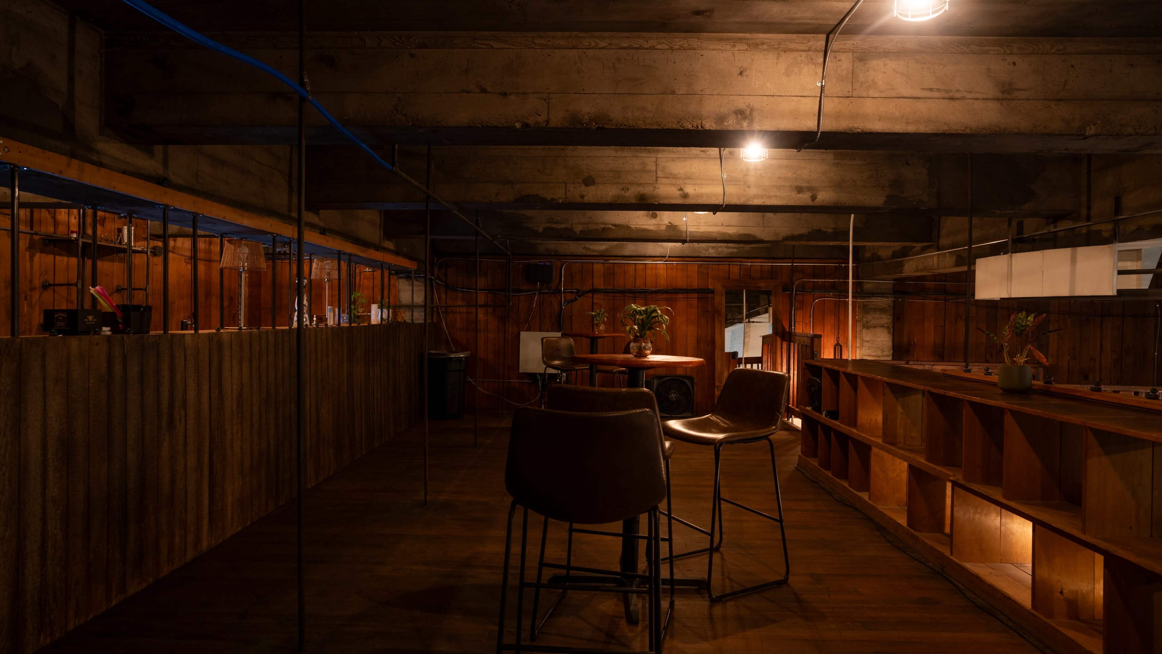 The image depicts a dimly lit, industrial-style interior featuring a wooden bar, a few high chairs, and shelves against the walls.