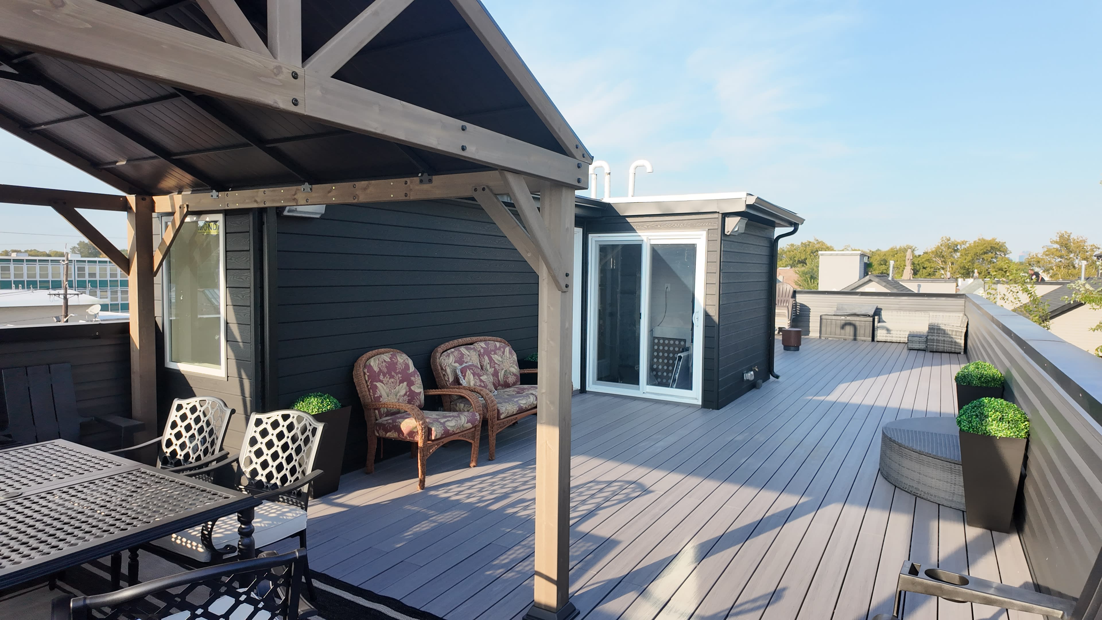 The image shows a rooftop terrace featuring a gazebo, outdoor seating, and potted plants, with a clear blue sky in the background.