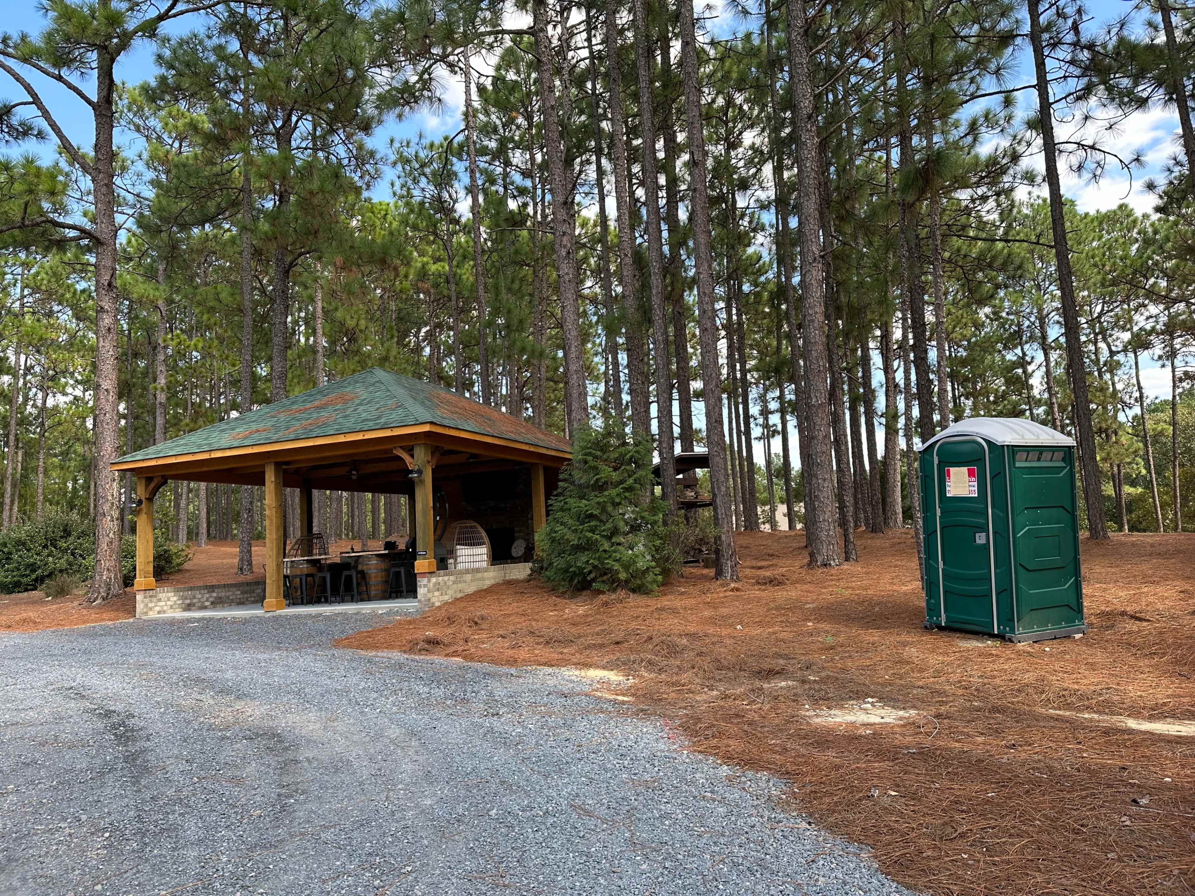 A wooden gazebo with a green roof is located beside a portable restroom in a wooded area filled with pine trees.