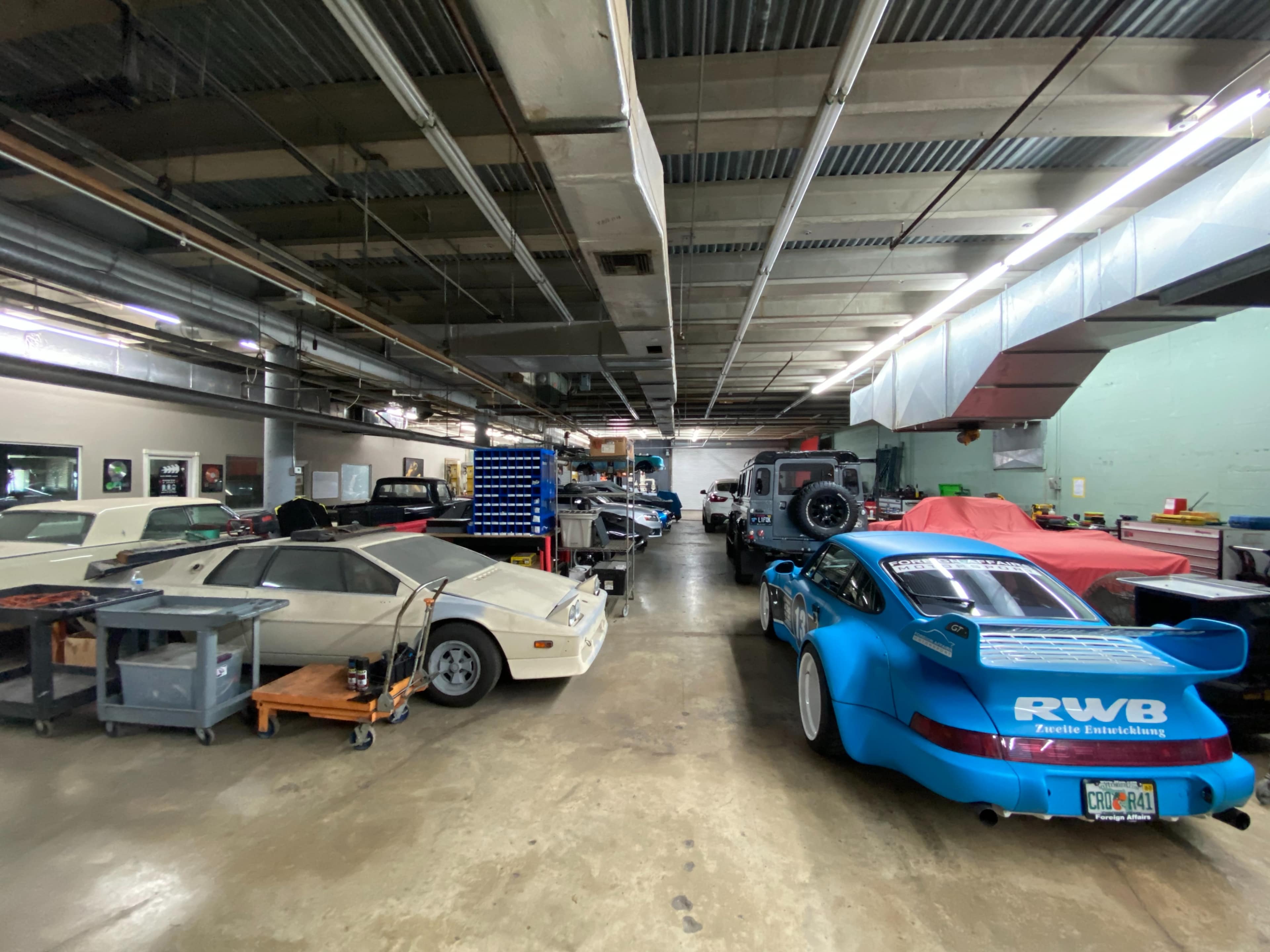 The image shows a garage filled with several cars, including a blue Porsche and a white sports car, with various tools and workstations along the sides.