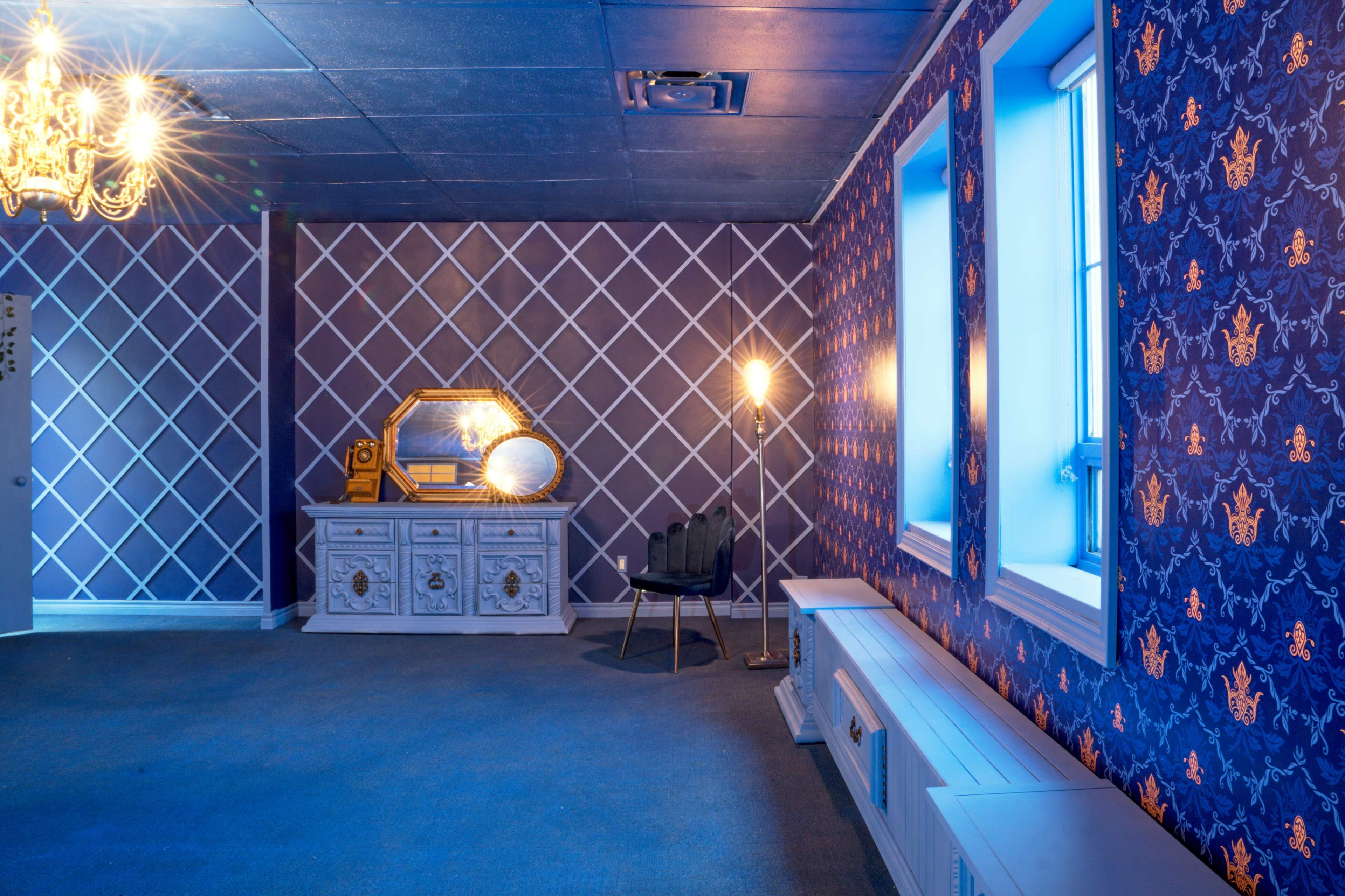 The room features a patterned wallpaper with a chandelier, a decorative mirror on a dresser, and a chair beside a lamp.