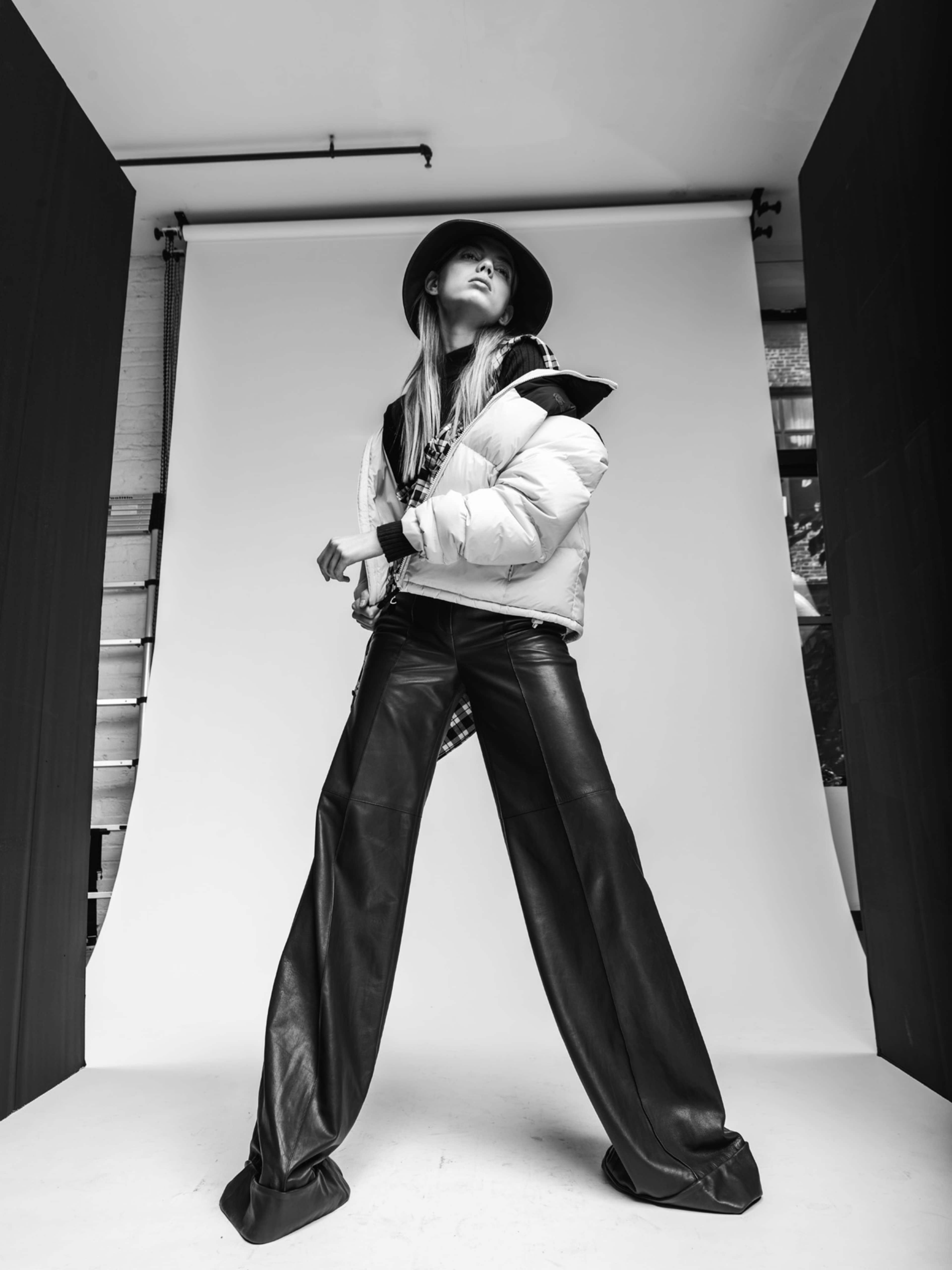 A model stands confidently in a studio, wearing a puffy jacket and wide-leg leather pants against a neutral backdrop.