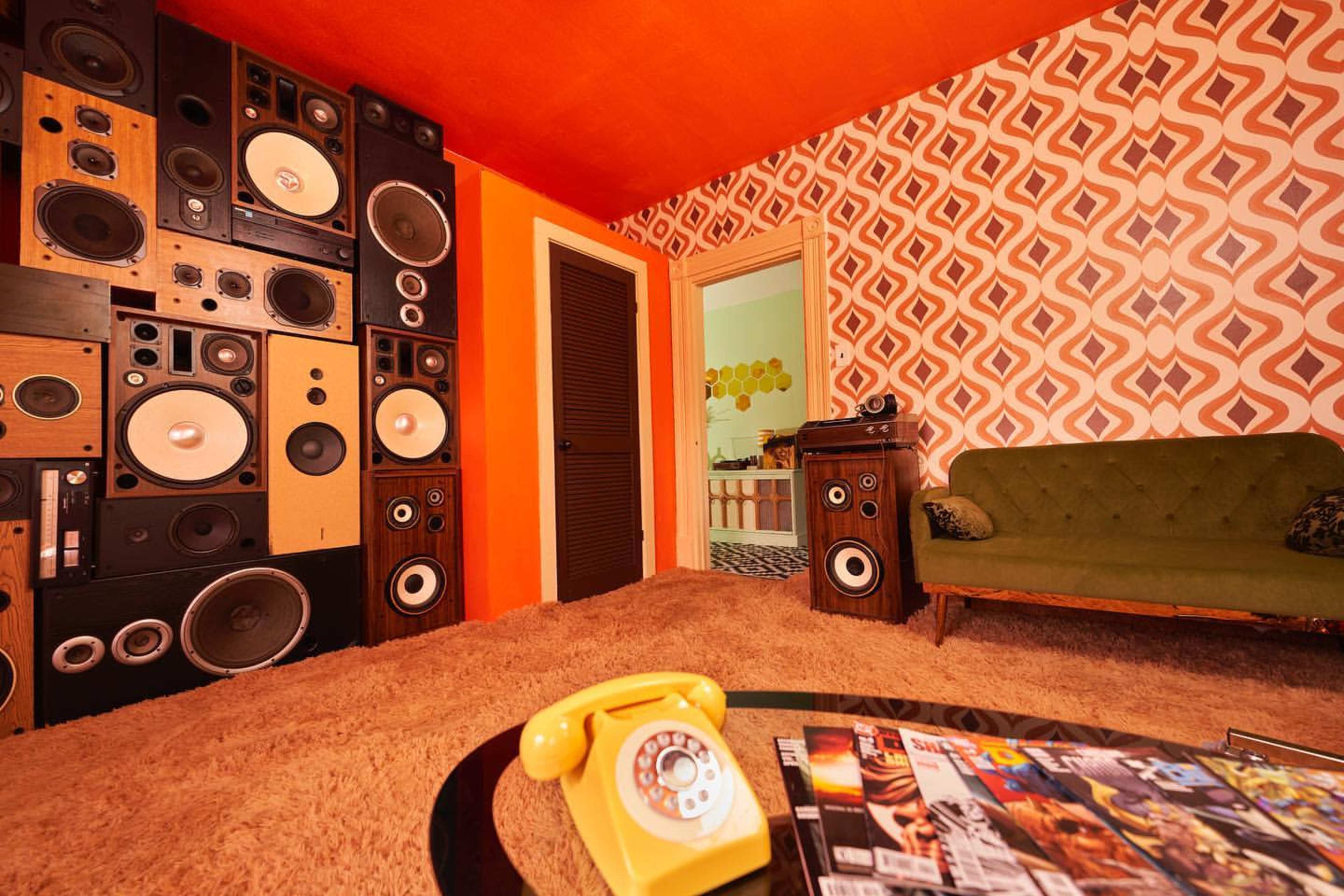 A retro-styled room features a wall of large speakers, an orange ceiling, and a green couch on a shag carpet with a yellow rotary phone and magazines on a side table.