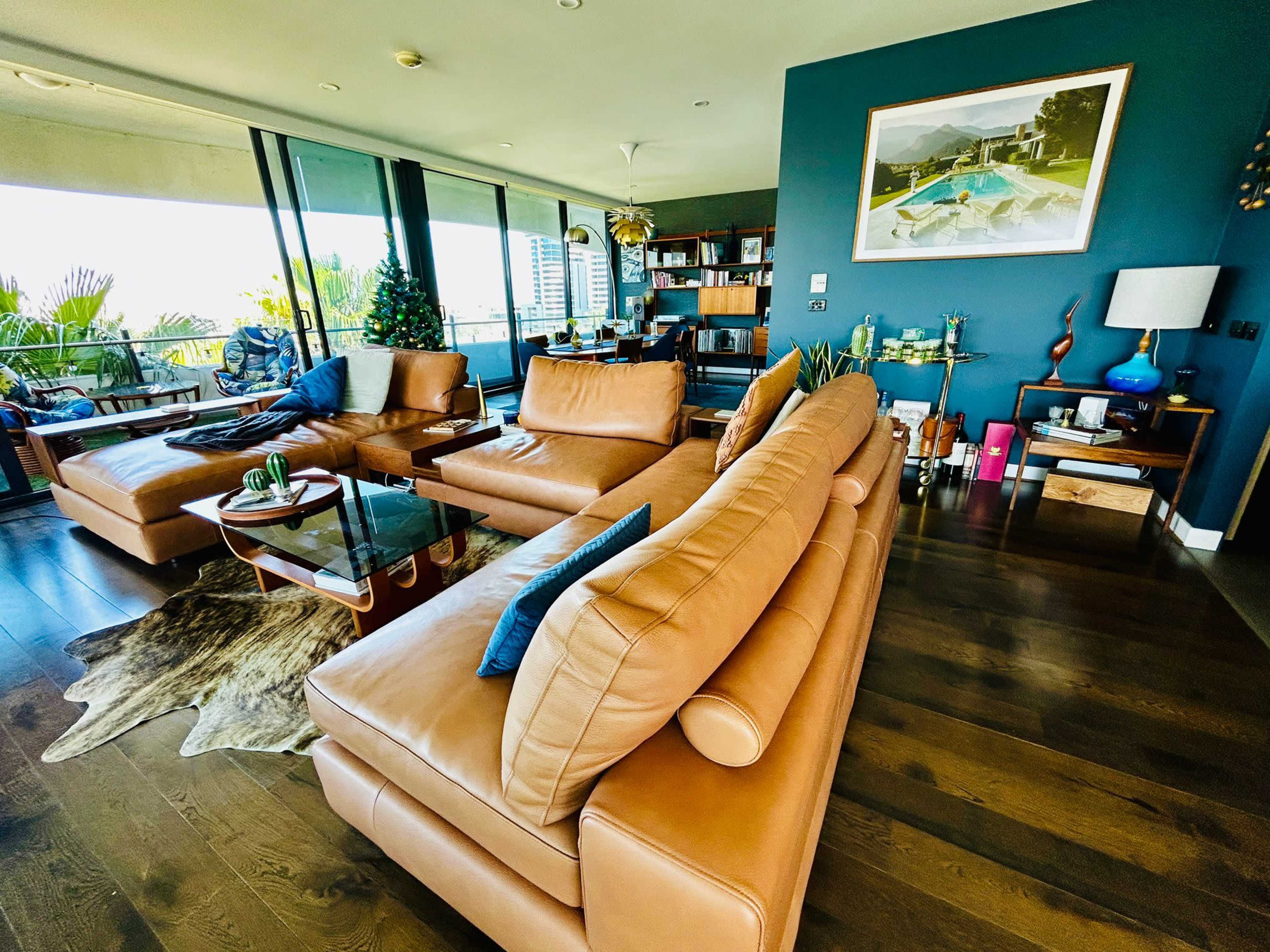 The image shows a modern living room with a large leather sectional sofa, a glass coffee table, and various decorative items on shelves against a dark blue wall.