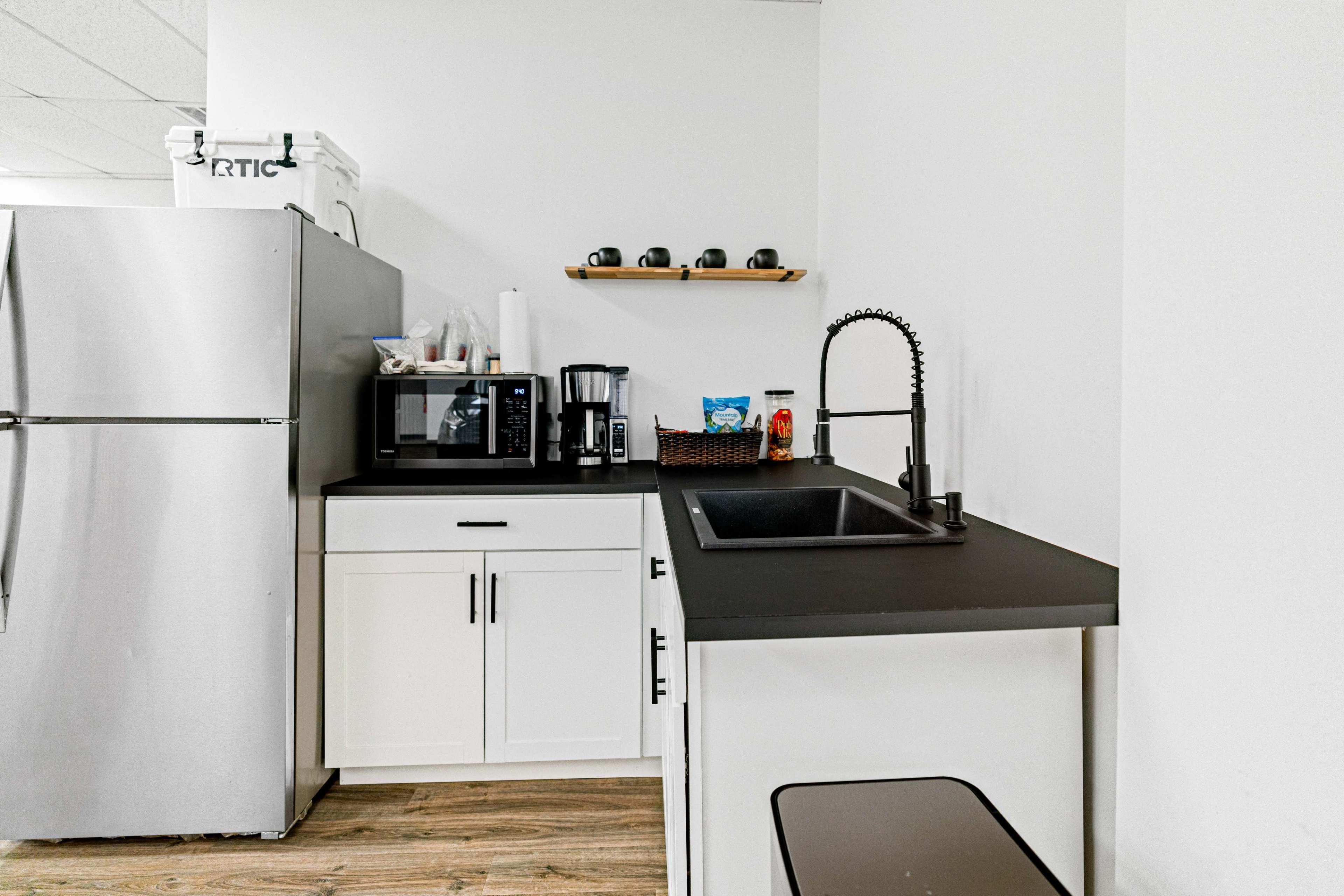 A modern kitchen corner with a stainless steel refrigerator, a microwave, a coffee maker, and a black sink on a countertop, all set against a white wall.